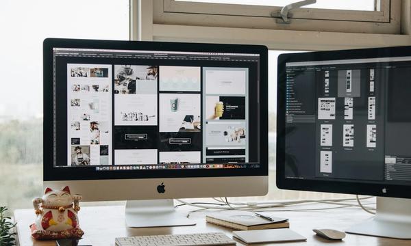 Two desktop monitors on a studio desk showing a grid of finished website page layouts, the kind of portfolio a web agency presents to a prospective client