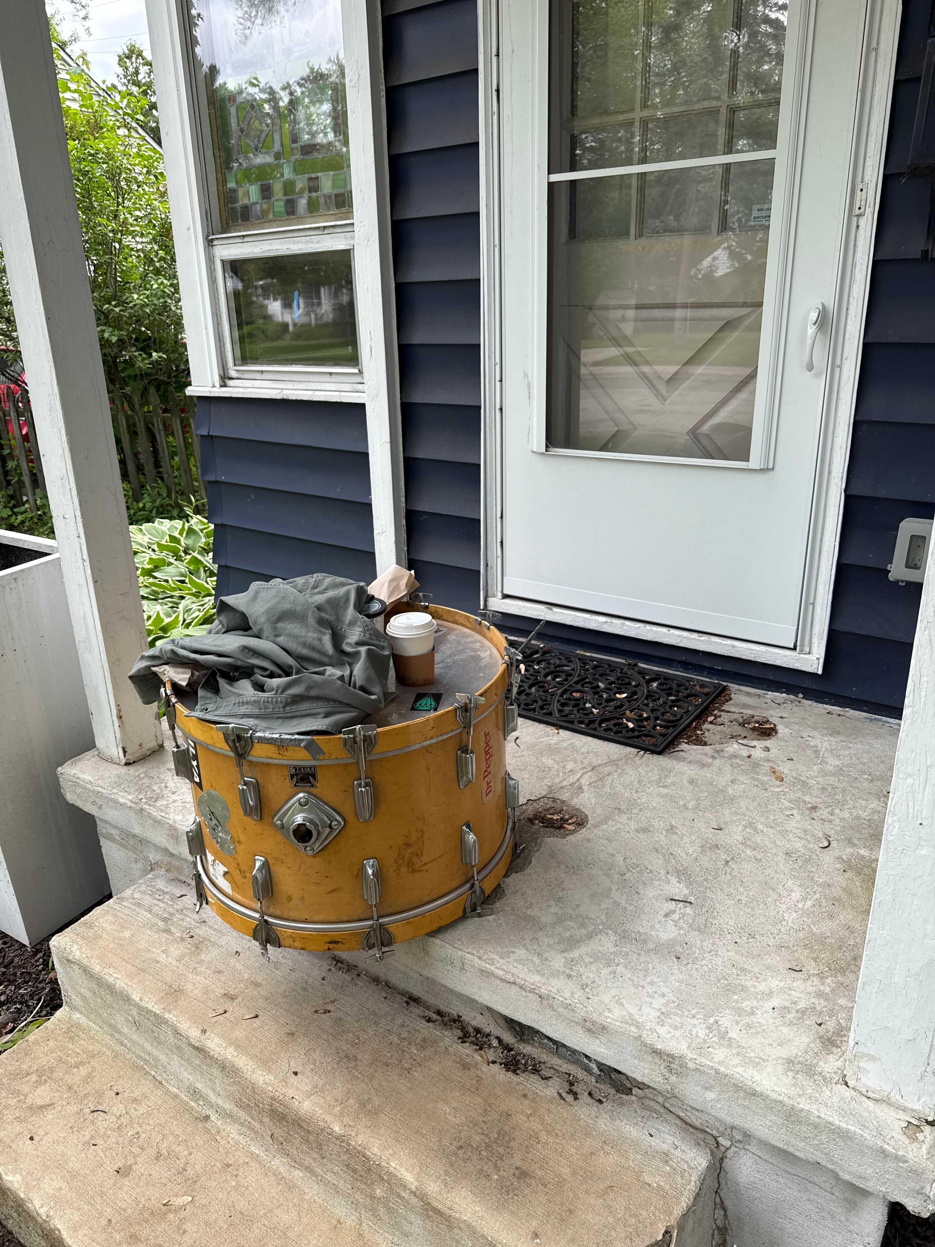 A kick drum on the porch of Fred's new blue house with white front door. A shirt and coffee cup is sitting on top on the kick drum. 