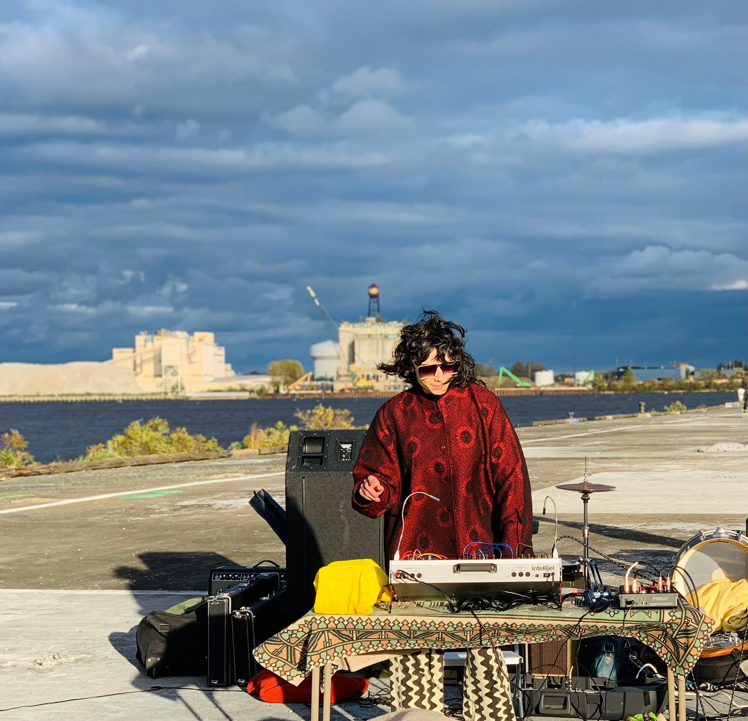 MANNA is performing outdoors. She has a modular synth riddled with patch cables perched on a blanketed tabletop. A larger speaker appears behind her. Off in the distance is a body of water. The sky seems both calm and ominous with pastel-gray clouds painted across the sky