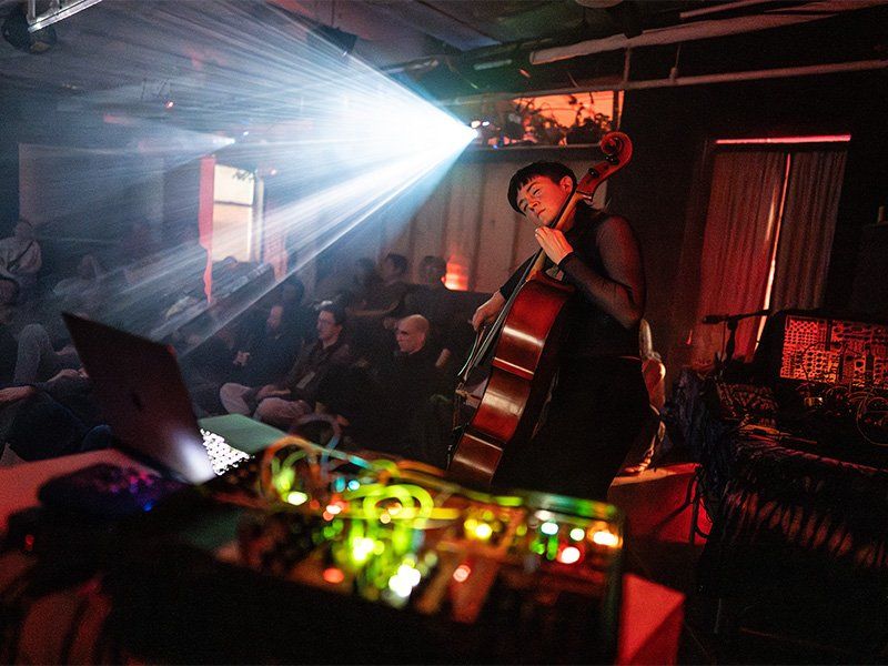 Kirin performs on stage, playing her cello, eyes closed, with a projector illuminating the audience behind her.