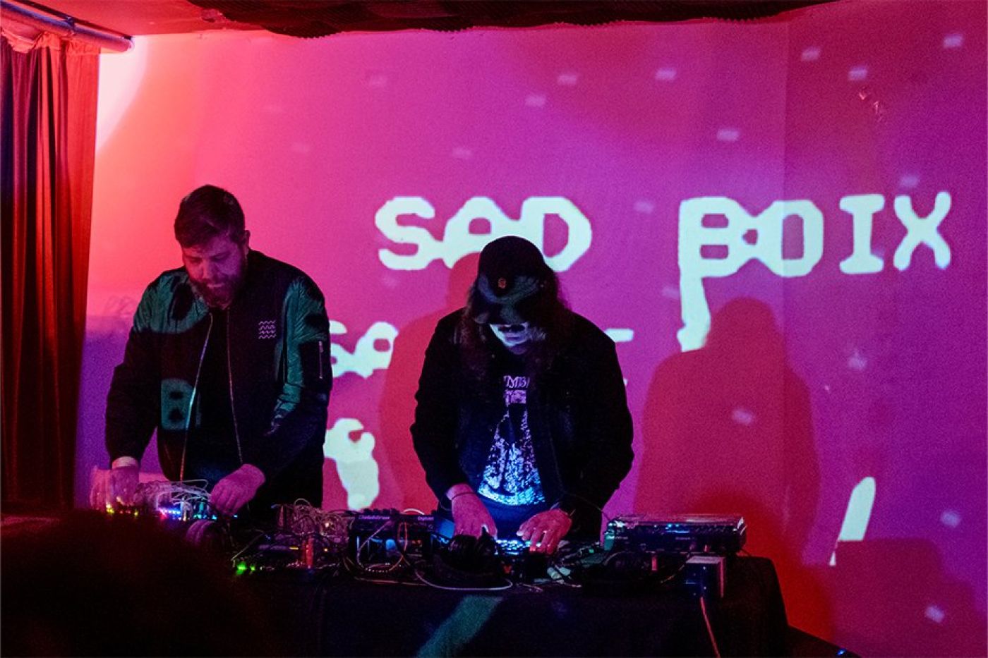 Two young men look down at a table filled with modular equipment, their hands fiddling with knobs and cables. Behind them is a projection that creates a deep pink hue in the room. The words "SAD BOIX" appear in an old arcade-style font.