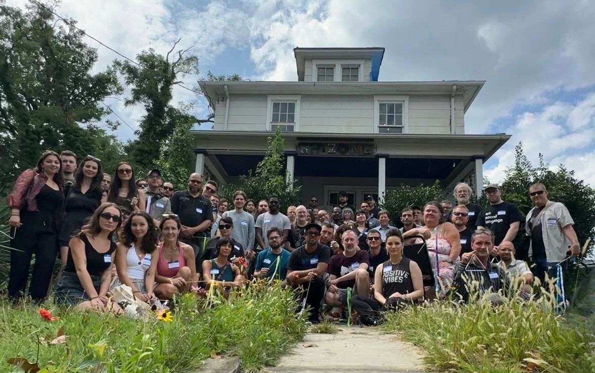 A crowd of folks come together to pose for a photo along the walkway which leads up to a large house behind them.