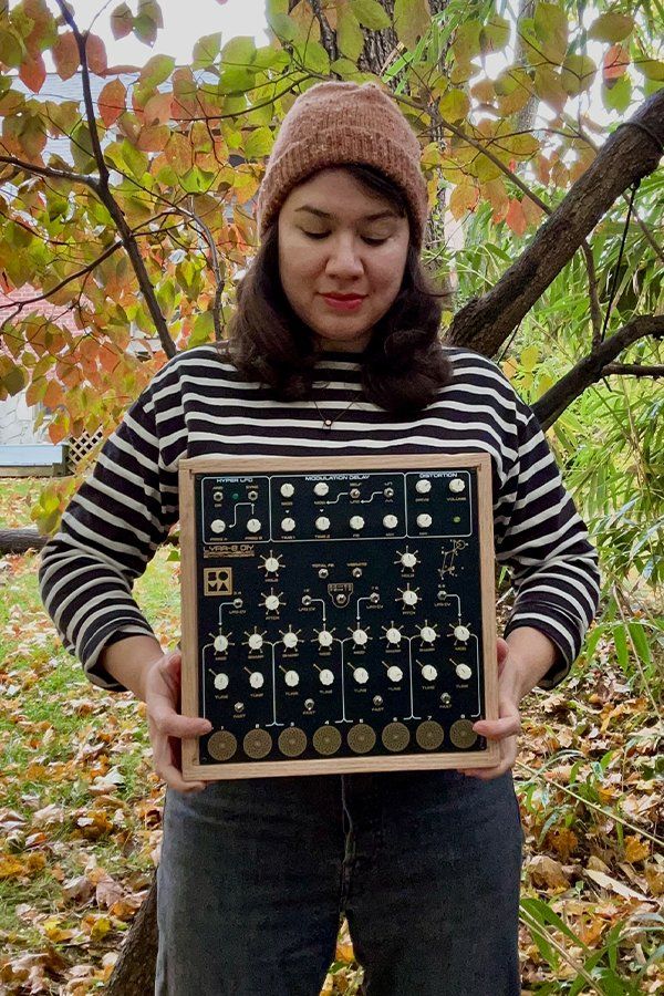 Carolyn is outdoors and holds up a modular synth unit for the camera.