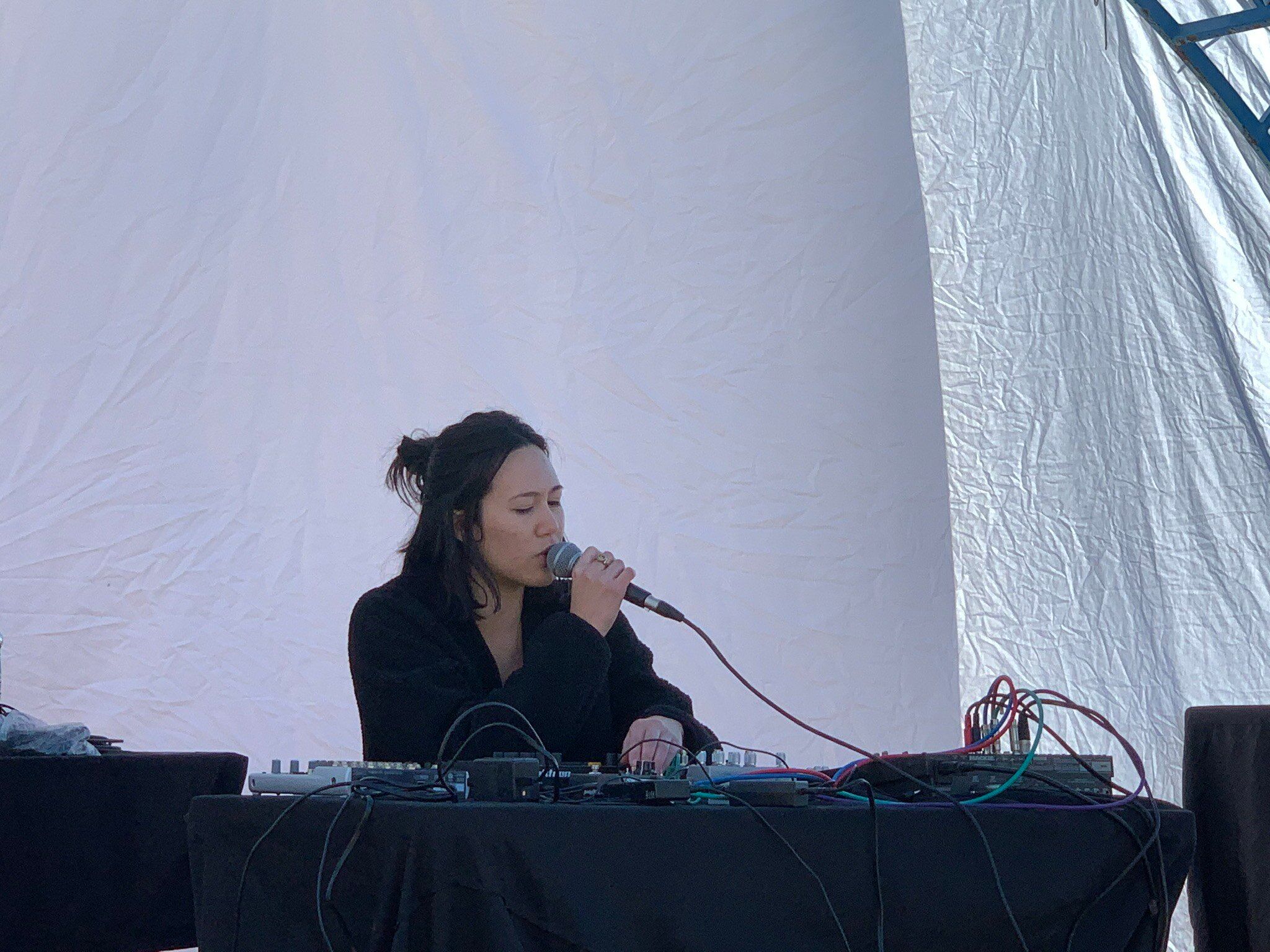 Emma is behind a table, singing into a microphone held in one hand. Her other hand is twisting knobs on a modular synth board placed on the table in front of her..