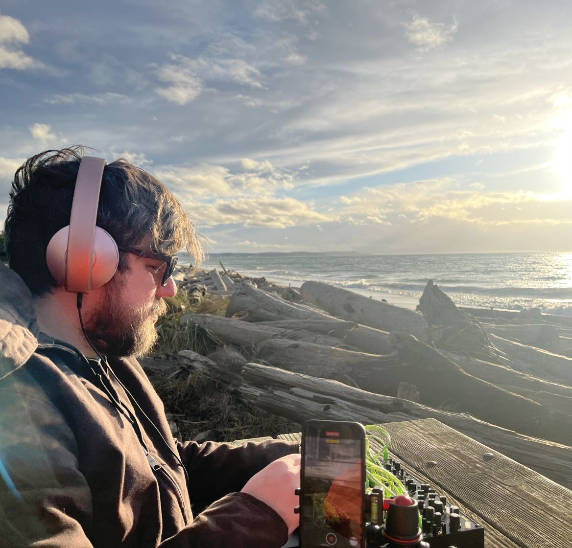 A close up shot of Toby's profile, him looking down at his modular synth. Behind him is a beautifully lush landscape, stratus clouds, calm ocean, and a large piece of driftwood. In the distance, the sun is beginning to set.
