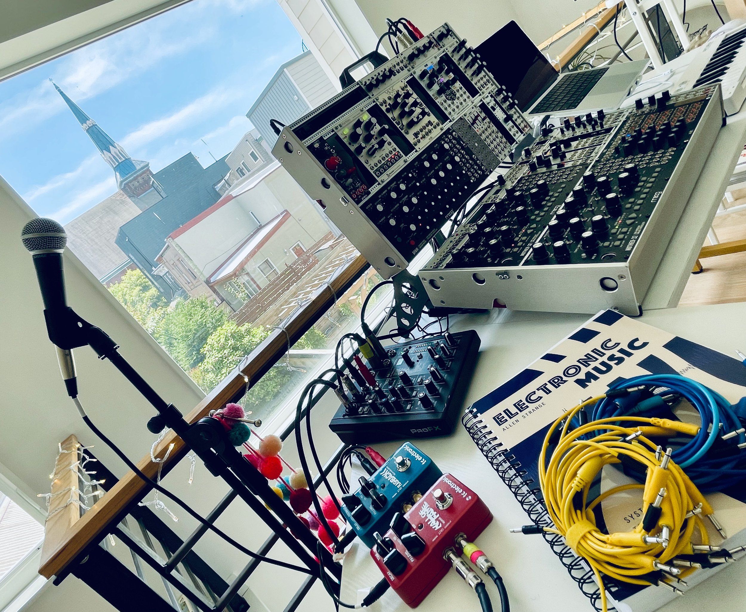 A desk covered in tons of excellent gear from a modular synth to effect pedals, cables, and a spiral-bound book with a cover that reads "Electronic Music"