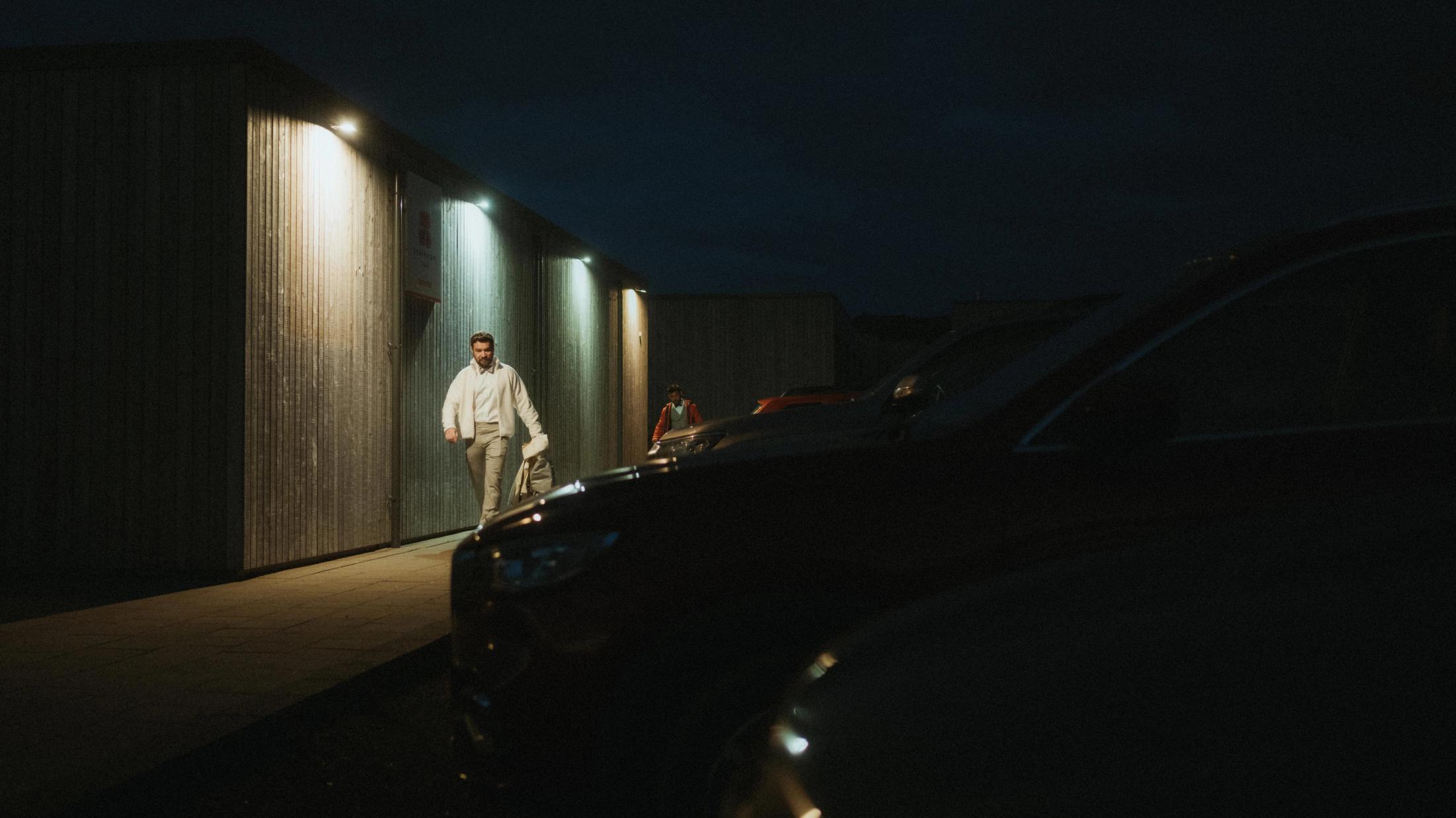 Ilya geht vor Sonnenaufgang von einem beleuchteten Gebäude zu den geparkten Autos.