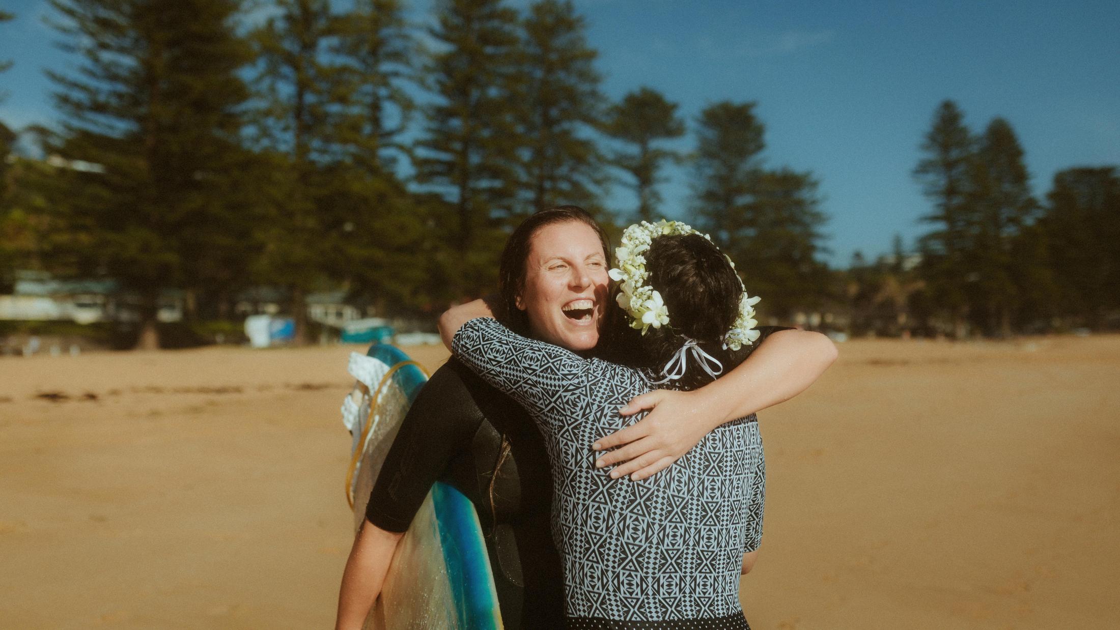 Petra laughing while embracing a woman carrying a surfboard