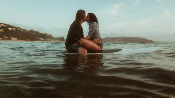 Petra and Roman kissing while seated together on a surfboard in the ocean.