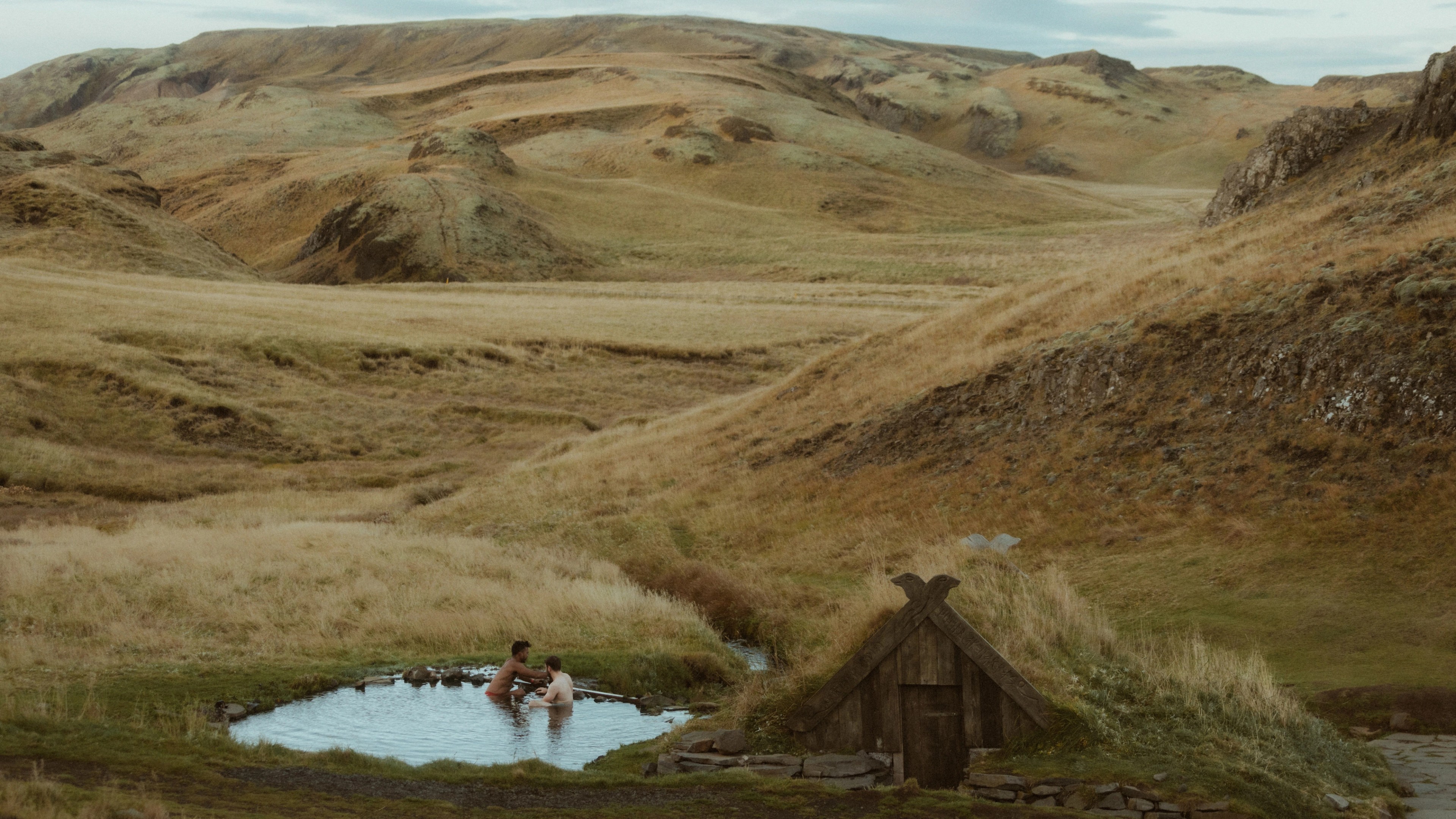 A tiny hot spring with Kris and Ilya sits beneath wide, empty Icelandic hills.