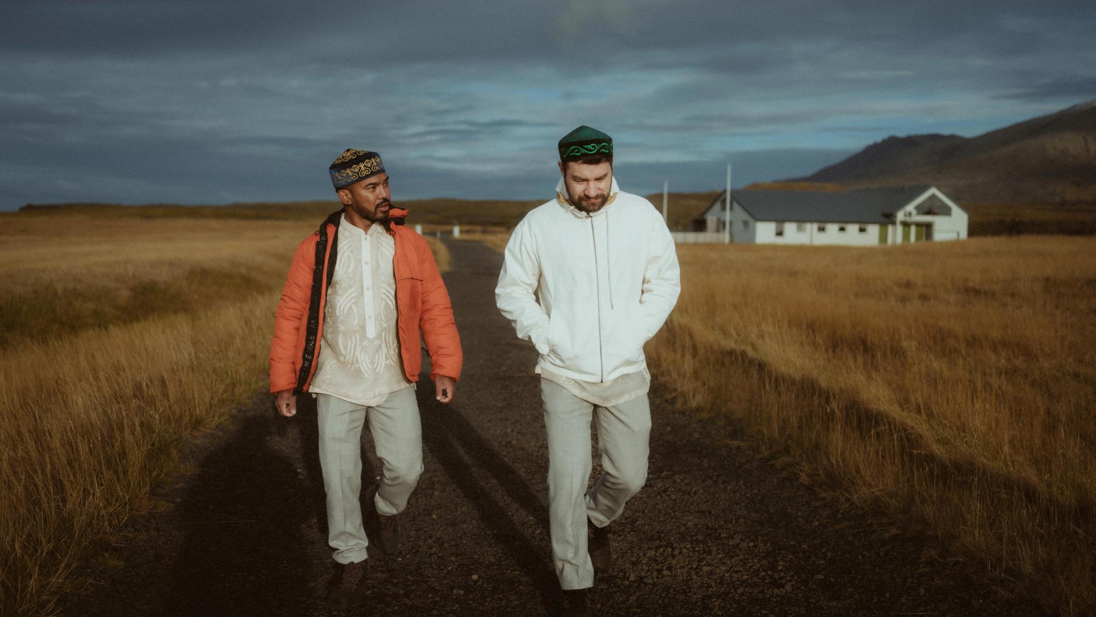 Kris und Ilya gehen in traditionellen Hemden und bestickten Kappen über ein offenes Feld.