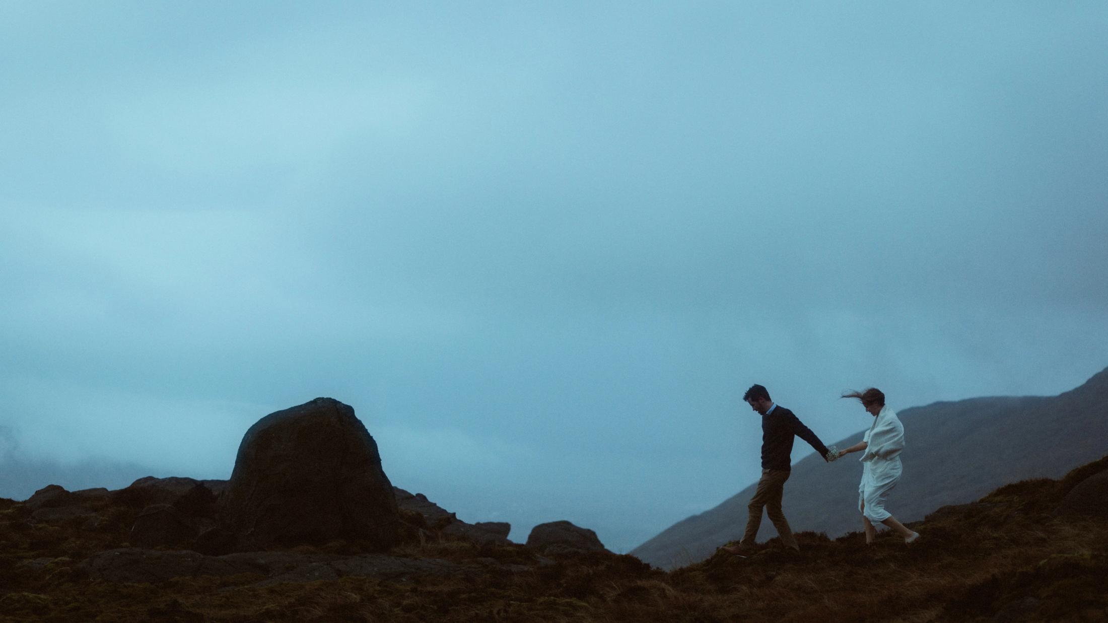 Jule und Rory gehen Hand in Hand über einen windigen Hang neben einem großen Felsen.
