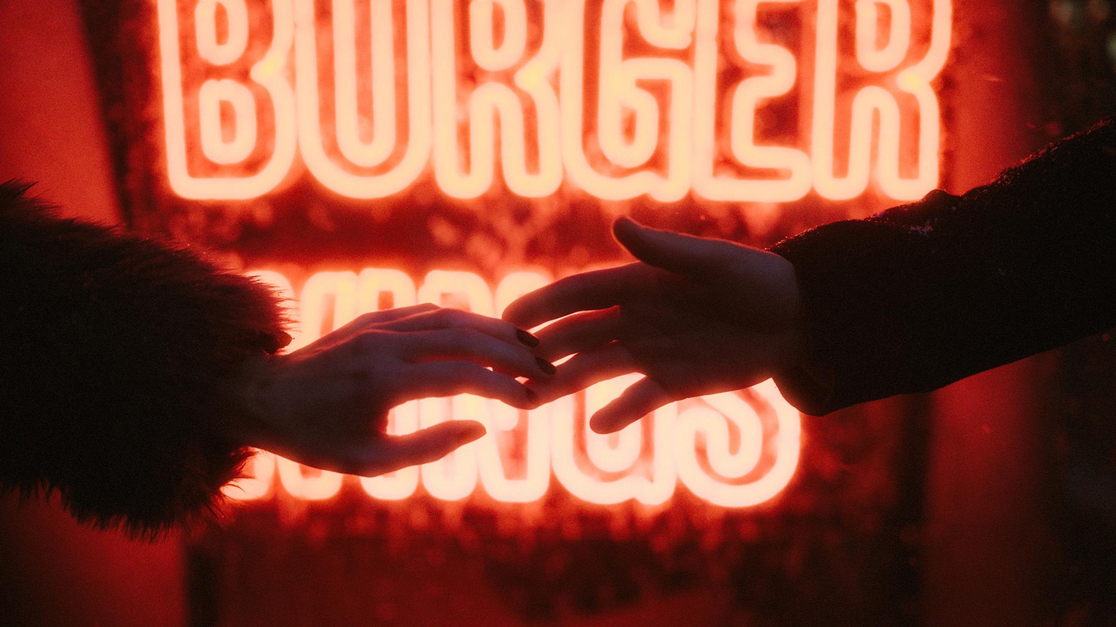 Lorelai's and Steffen's hands reach toward each other against red neon lettering.