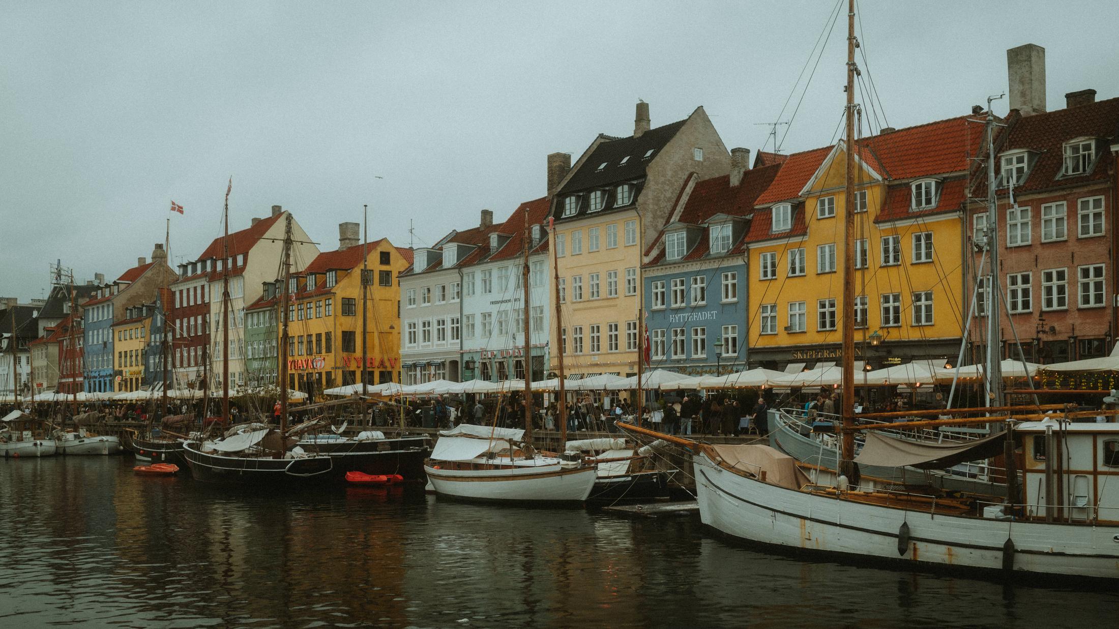 Boats sit below a row of colourful Nyhavn facades beneath a grey sky.