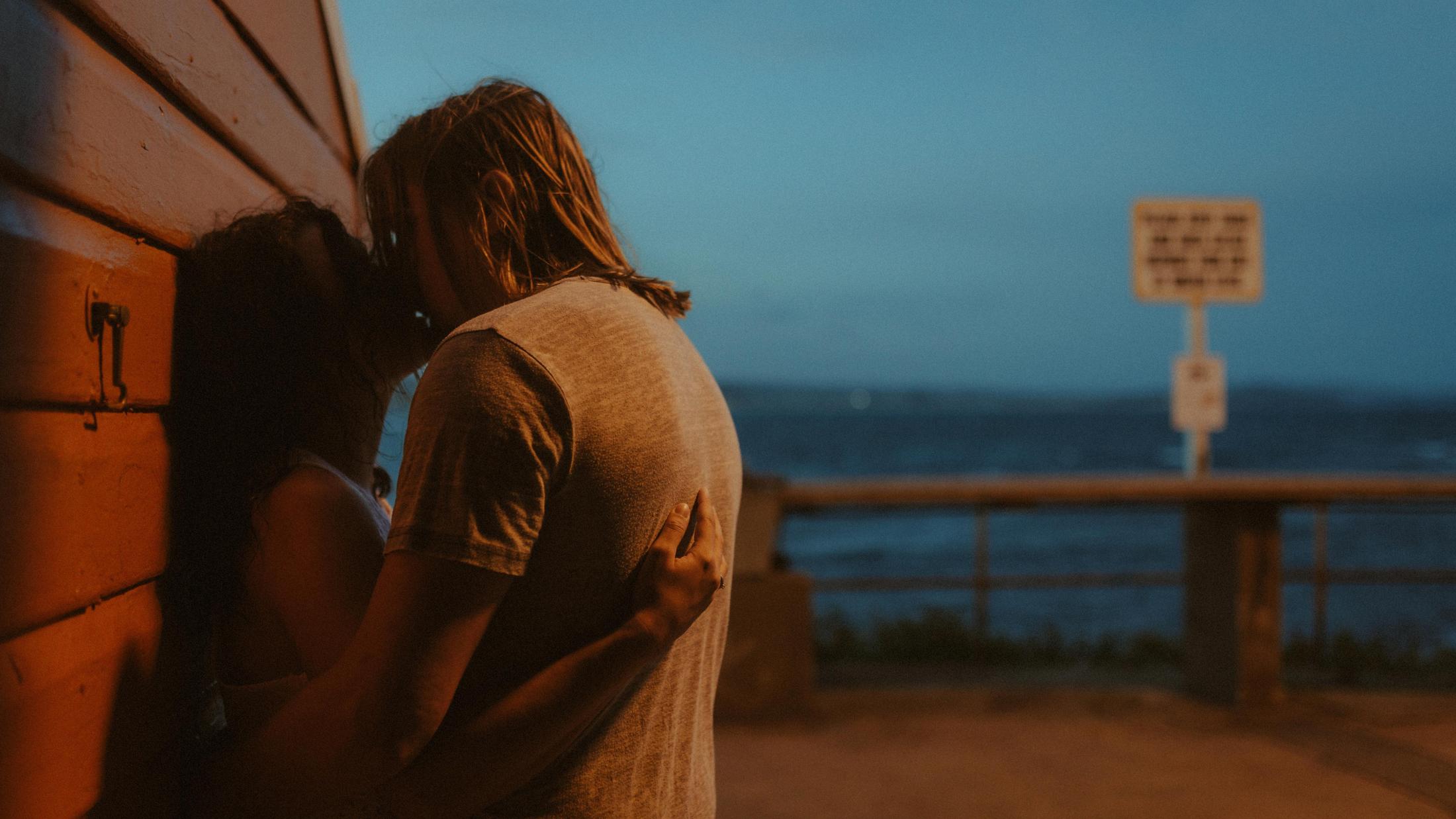 Petra and Roman kissing beside the boatshed at dusk