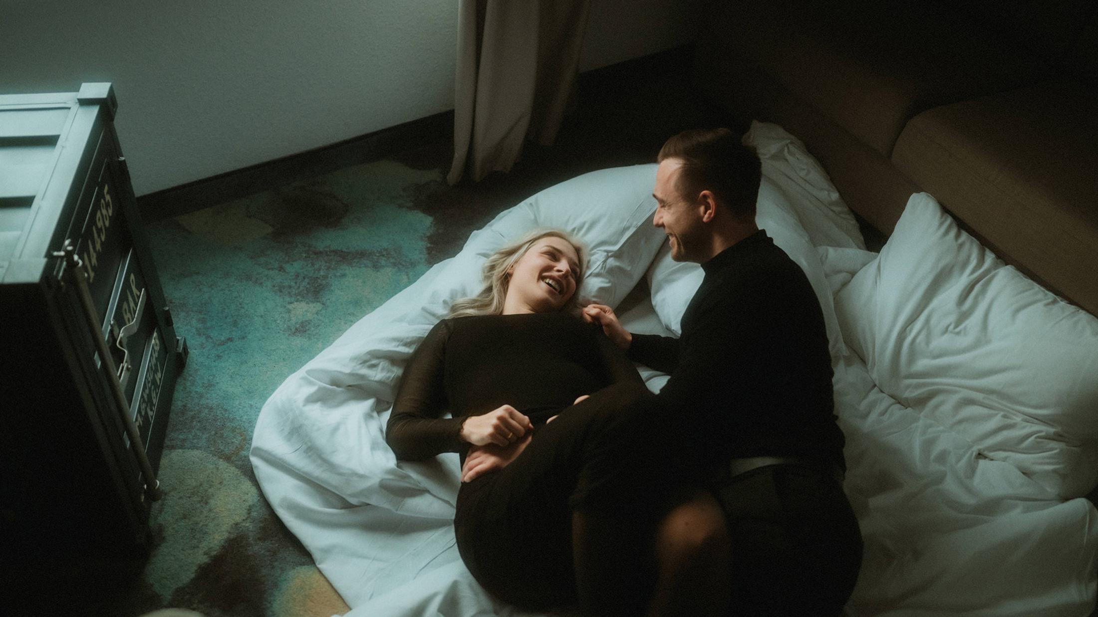 Lorelai laughs beside Steffen on an unmade hotel bed, seen from above.