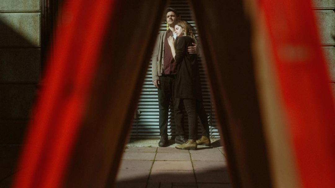 Aiste and John standing together in sunlight, seen through a blurred red foreground