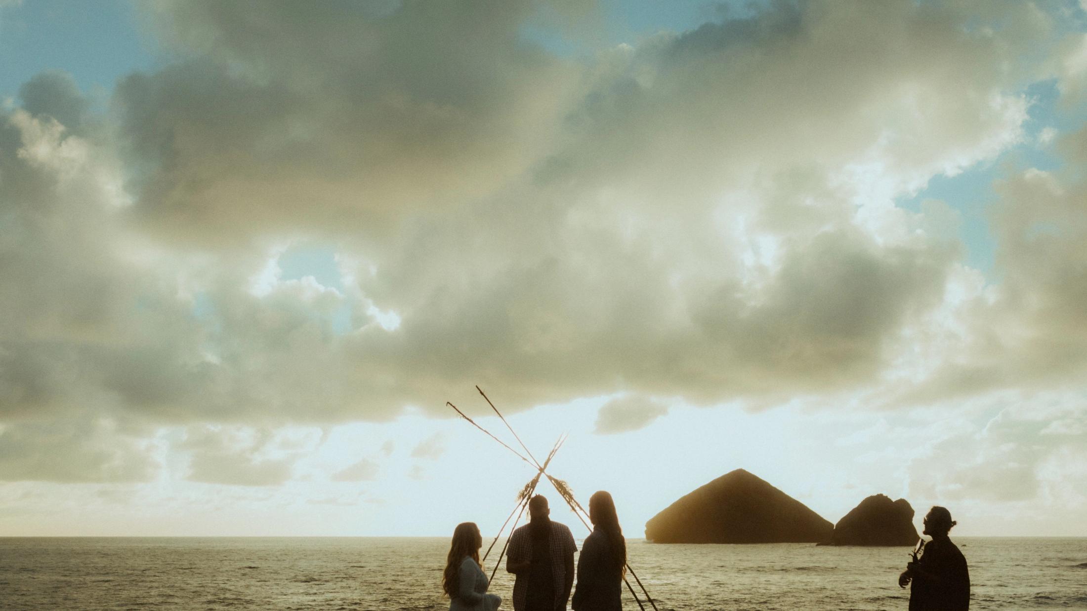 The couple and officiant appear small beneath a broad clouded sky at the beach.