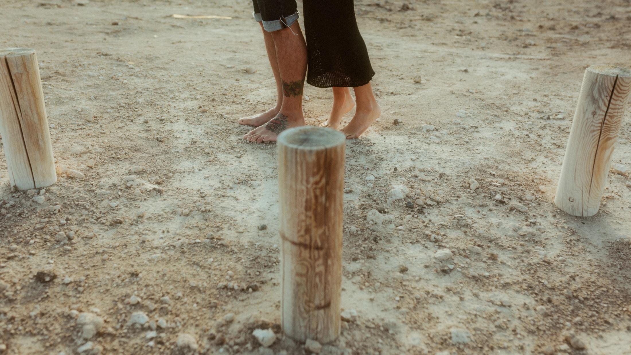 Holzpfähle im Vordergrund, die nackten Füße des Paares gehen auf einem staubigen Weg davon.