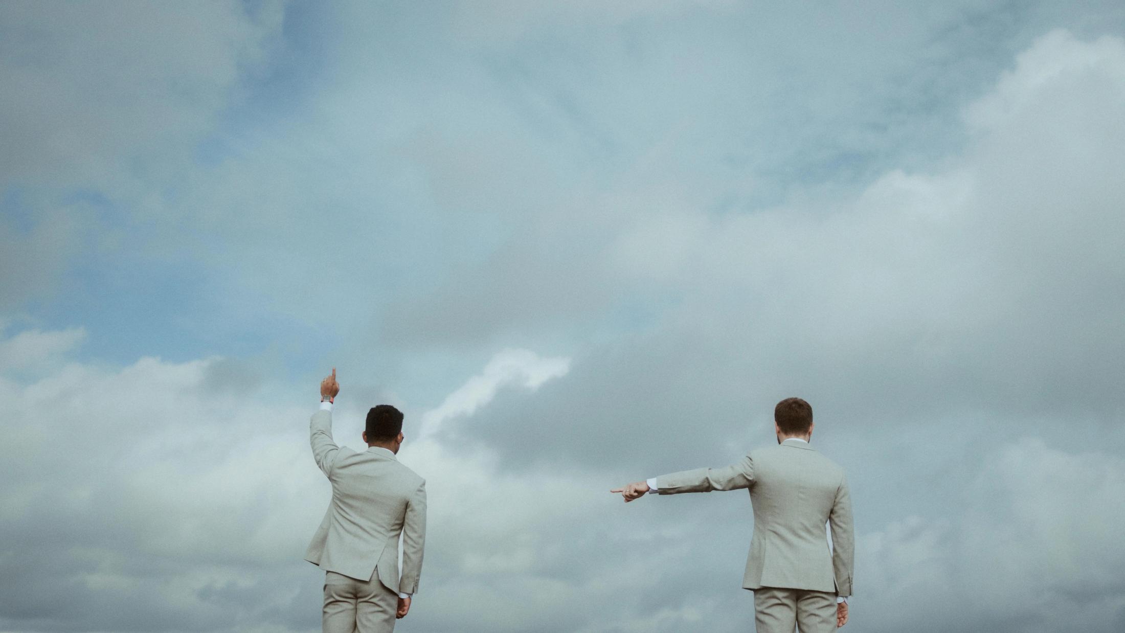 The couple stands beneath a cloudy sky with their arms extended in opposite directions.