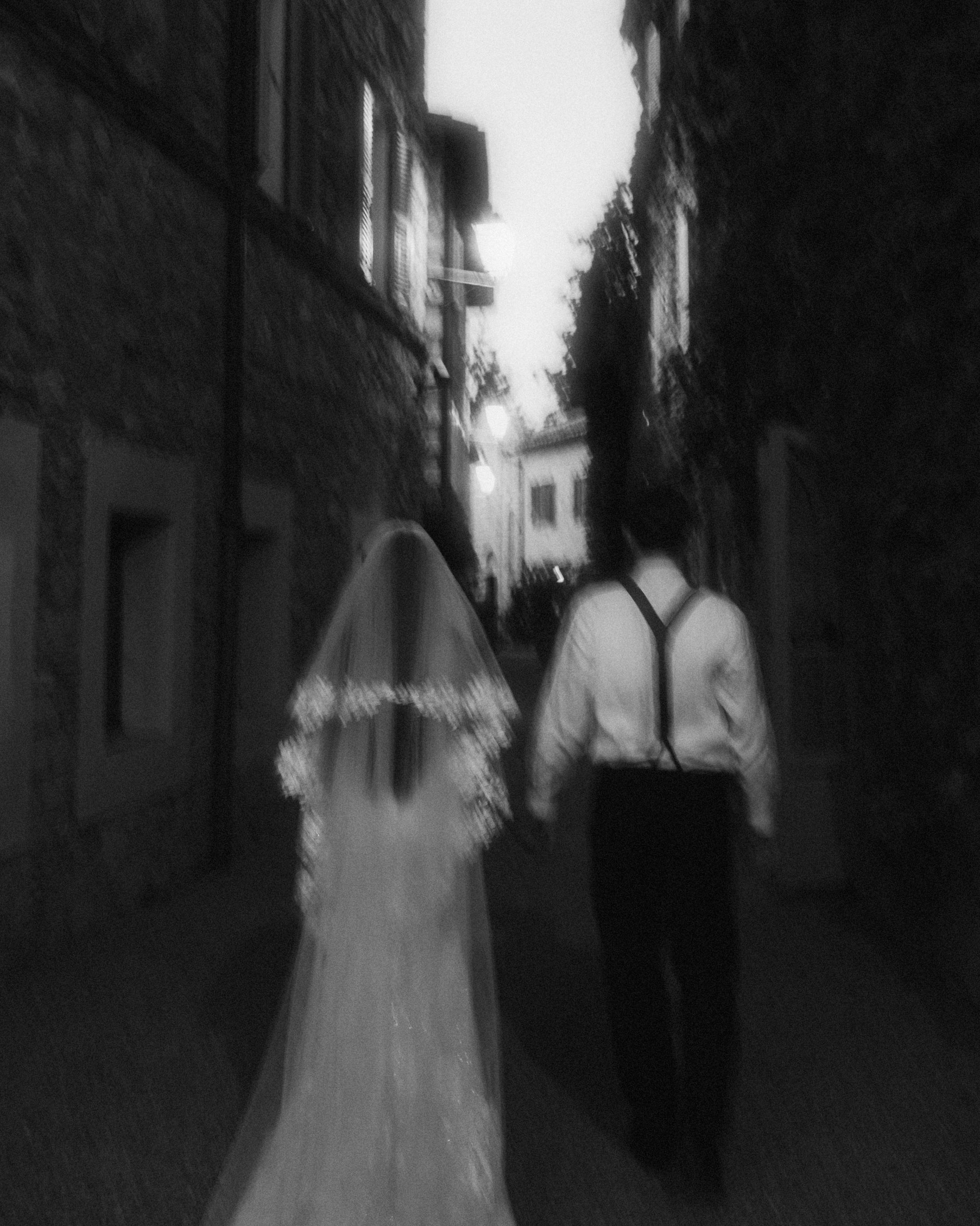 The couple walks away through a narrow shadowed lane in a blurred black-and-white frame.