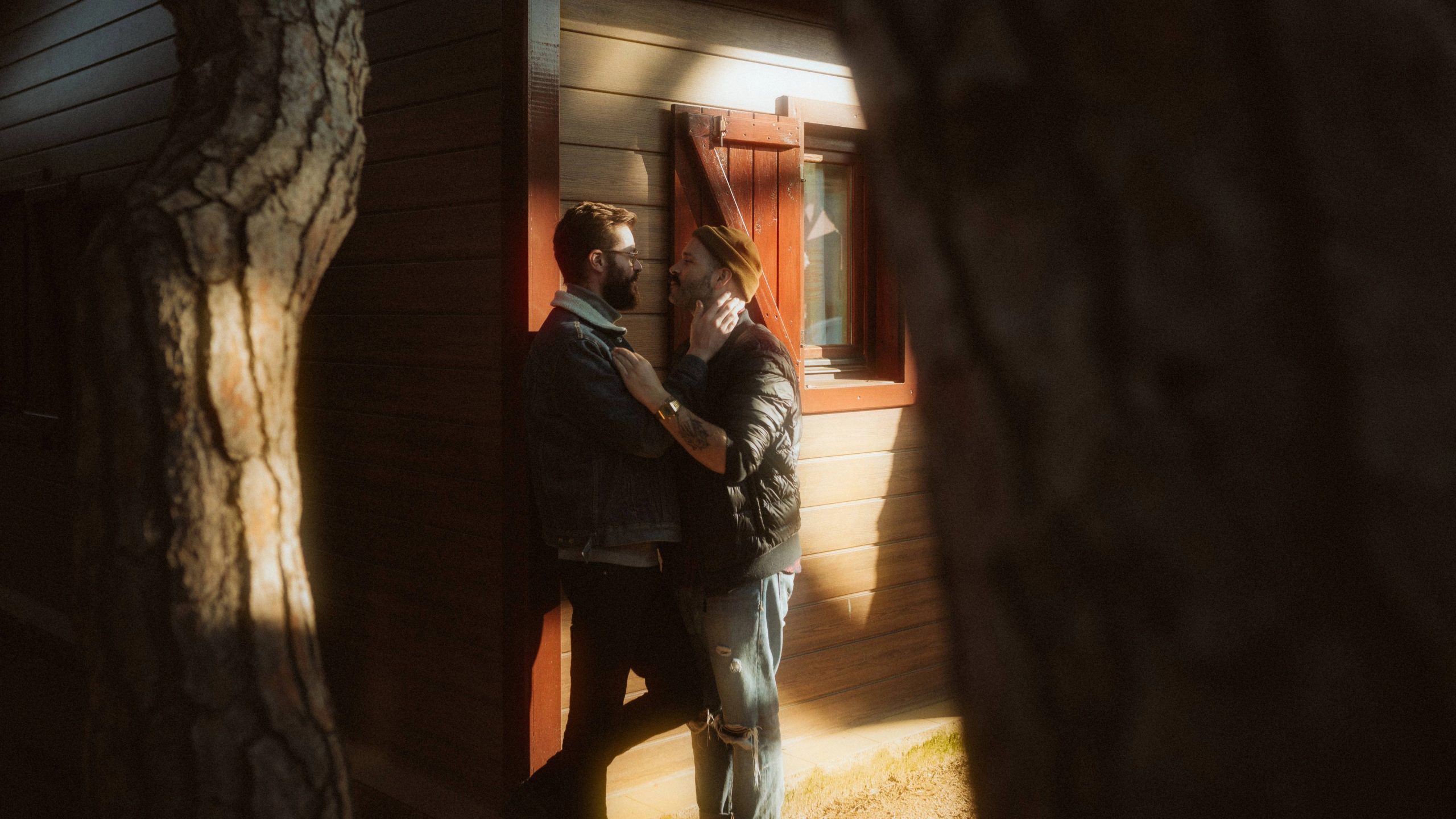 Two men in warm sunlight beside a wooden cabin during their lovers session