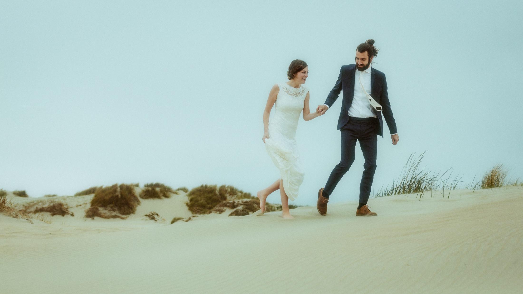 Nina und Torben gehen barfuß und Hand in Hand durch den Sand und schauen einander an.