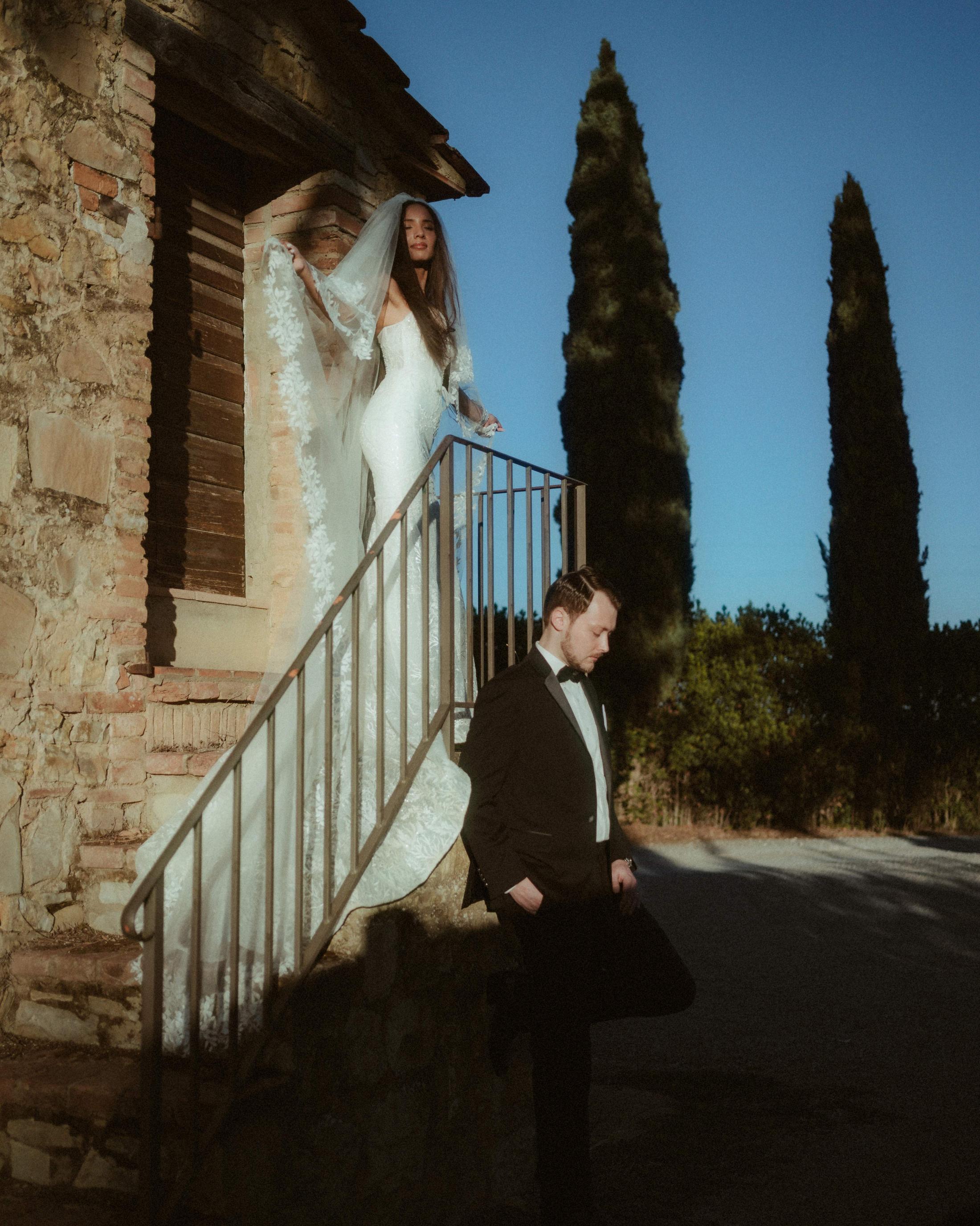 Vertical portrait of the couple on stone stairs beside tall cypress trees.