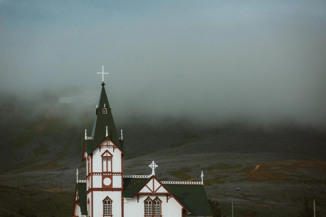 The red and white church tower in Húsavík with fog covering the hillside behind it.
