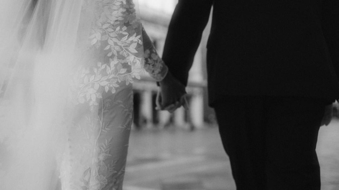 Black-and-white detail of joined hands beside the bride's lace dress.