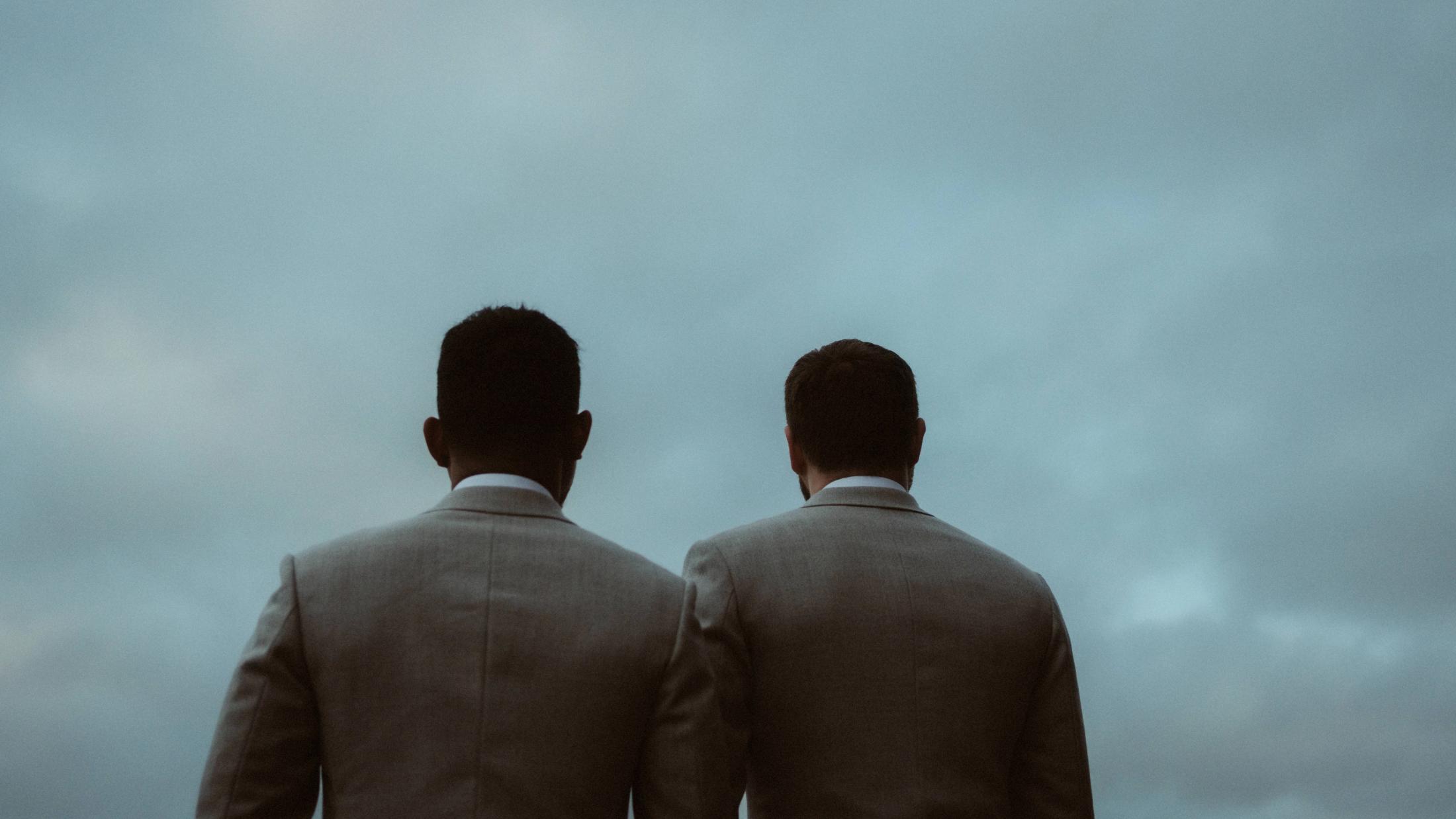 Kris and Ilya stand shoulder to shoulder with their backs to the camera beneath a wide grey sky.