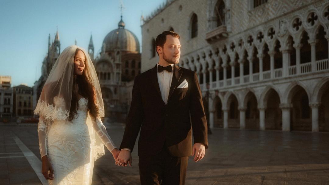 A couple walks hand in hand through warm morning light in St Mark’s Square in Venice.