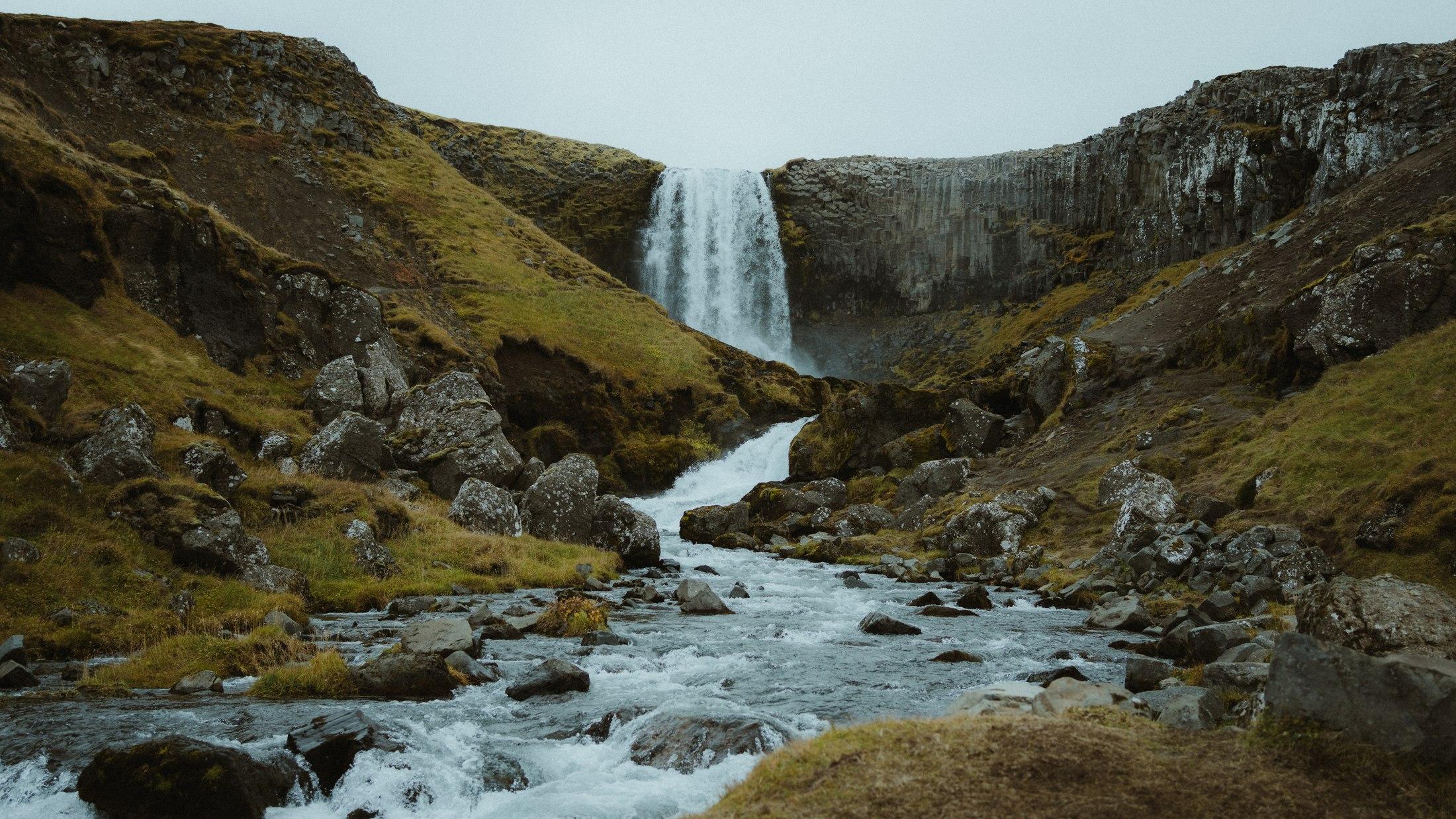 A broad waterfall falling over a basalt cliff, with a rocky stream winding through mossy ground.
