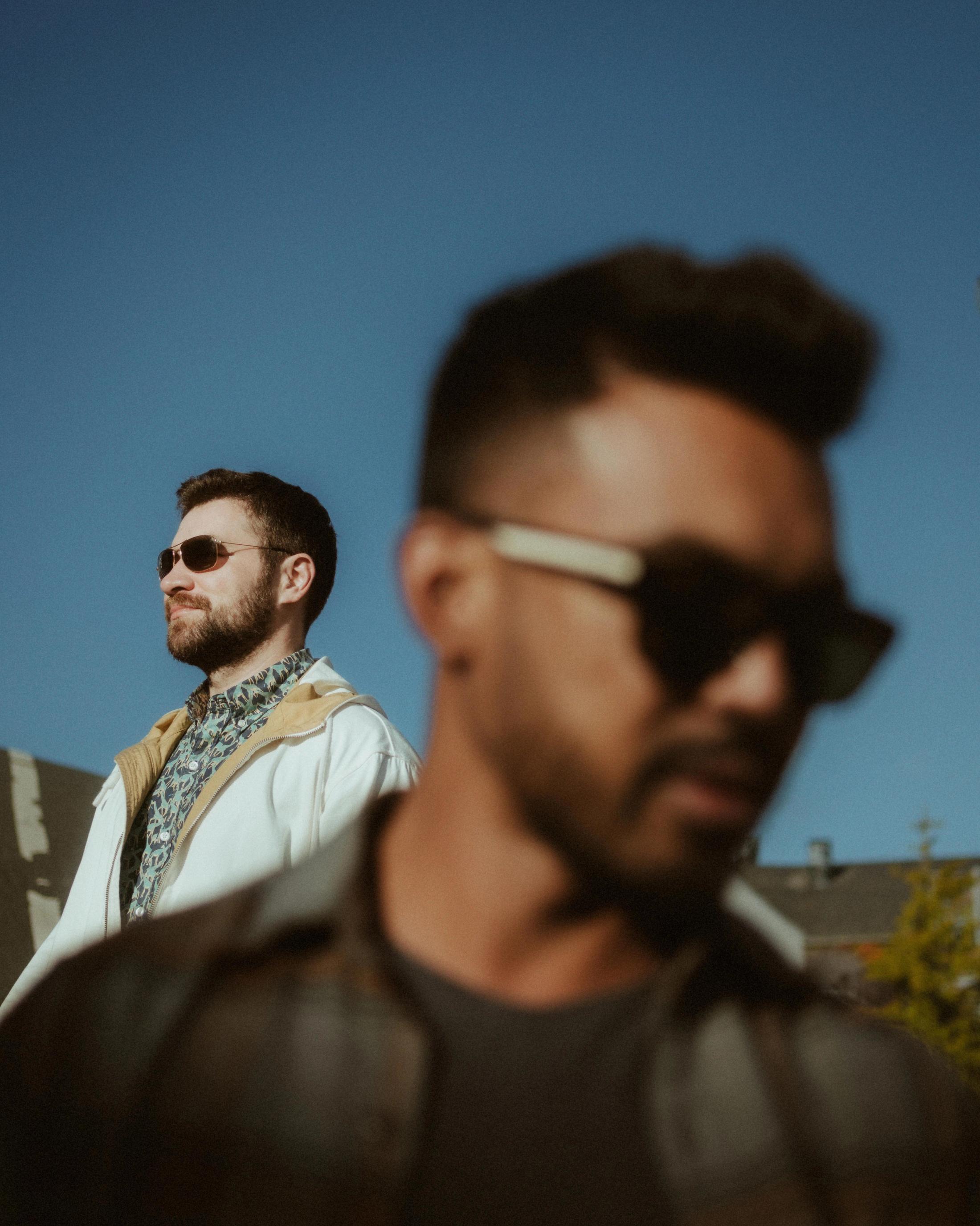Layered portrait of Kris and Ilya standing at different distances against a clear blue sky.