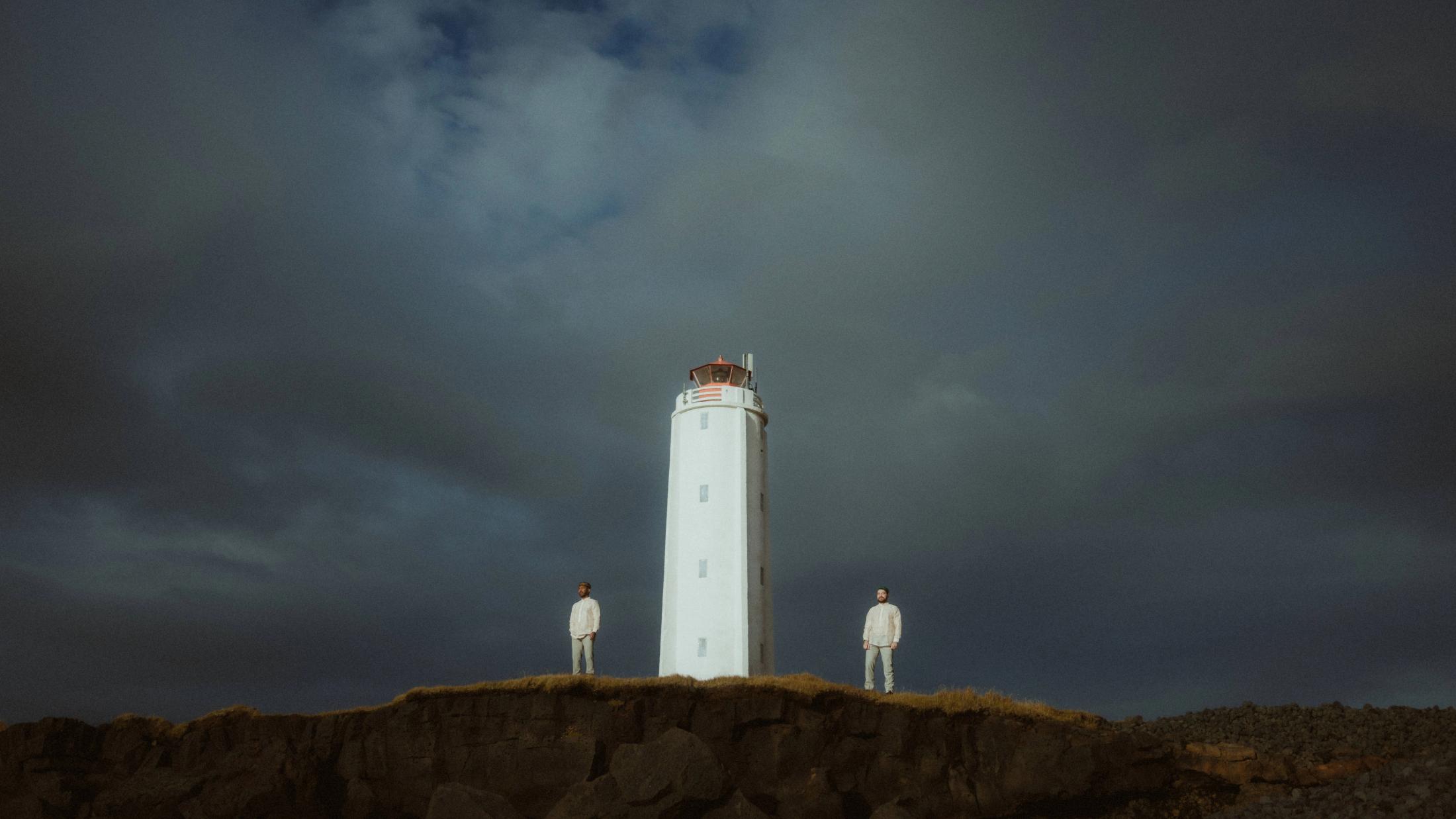 Kris und Ilya stehen als kleine Figuren unter einem weißen Leuchtturm.