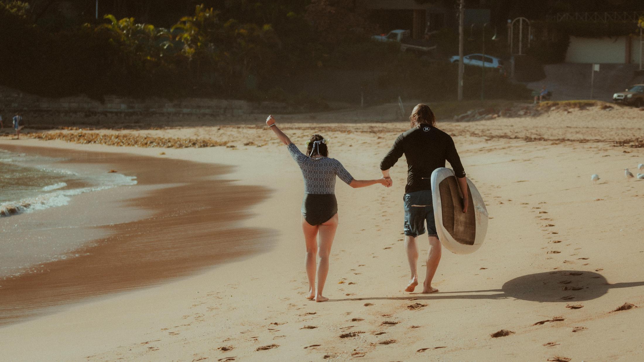 Petra and Roman walking hand in hand across the sand with a surfboard