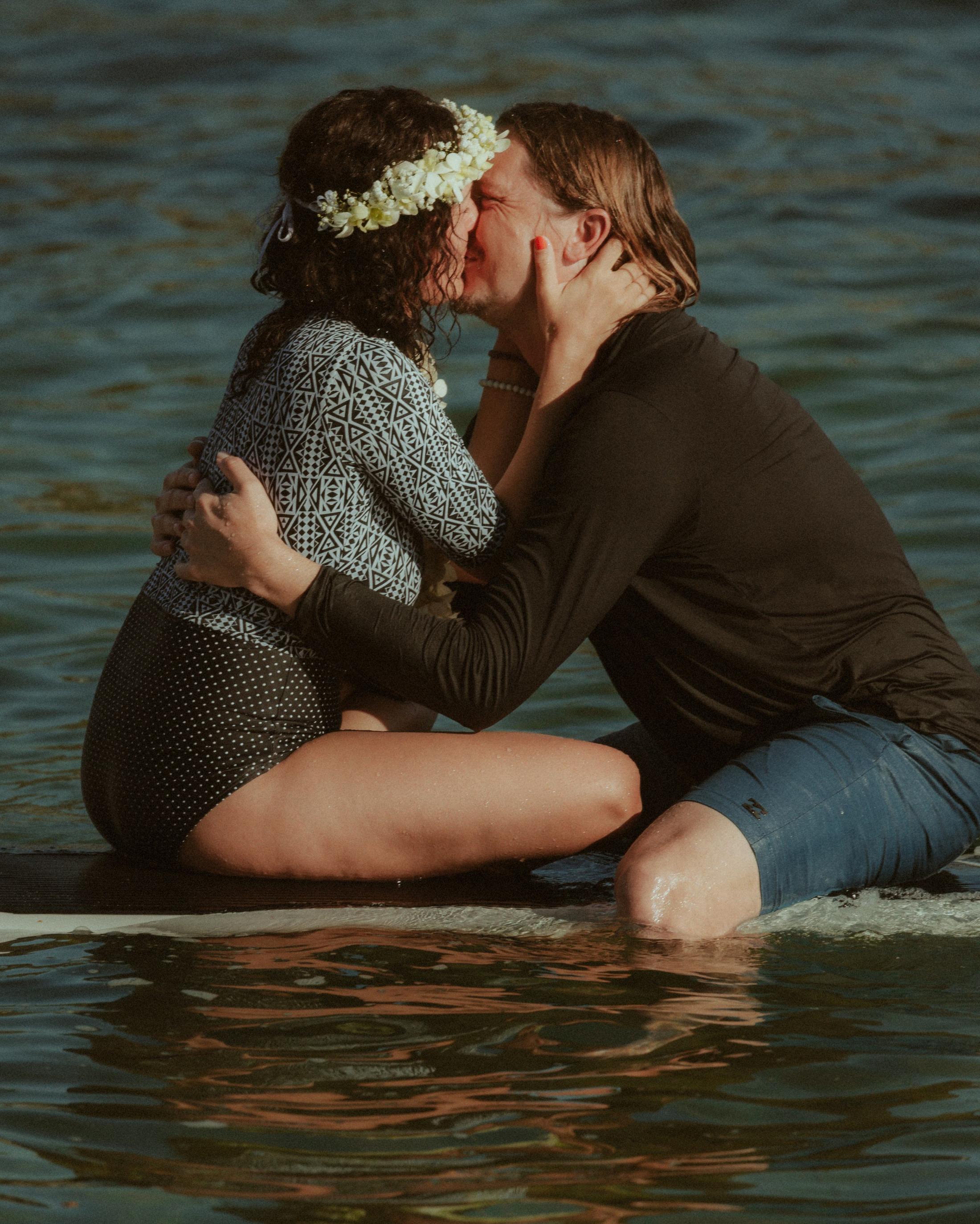 Petra and Roman kissing while seated together on a surfboard