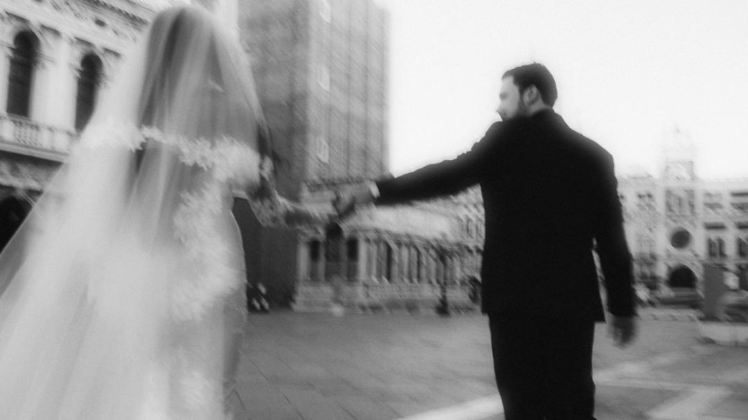 In a blurred black-and-white frame, the groom reaches for the bride's hand.