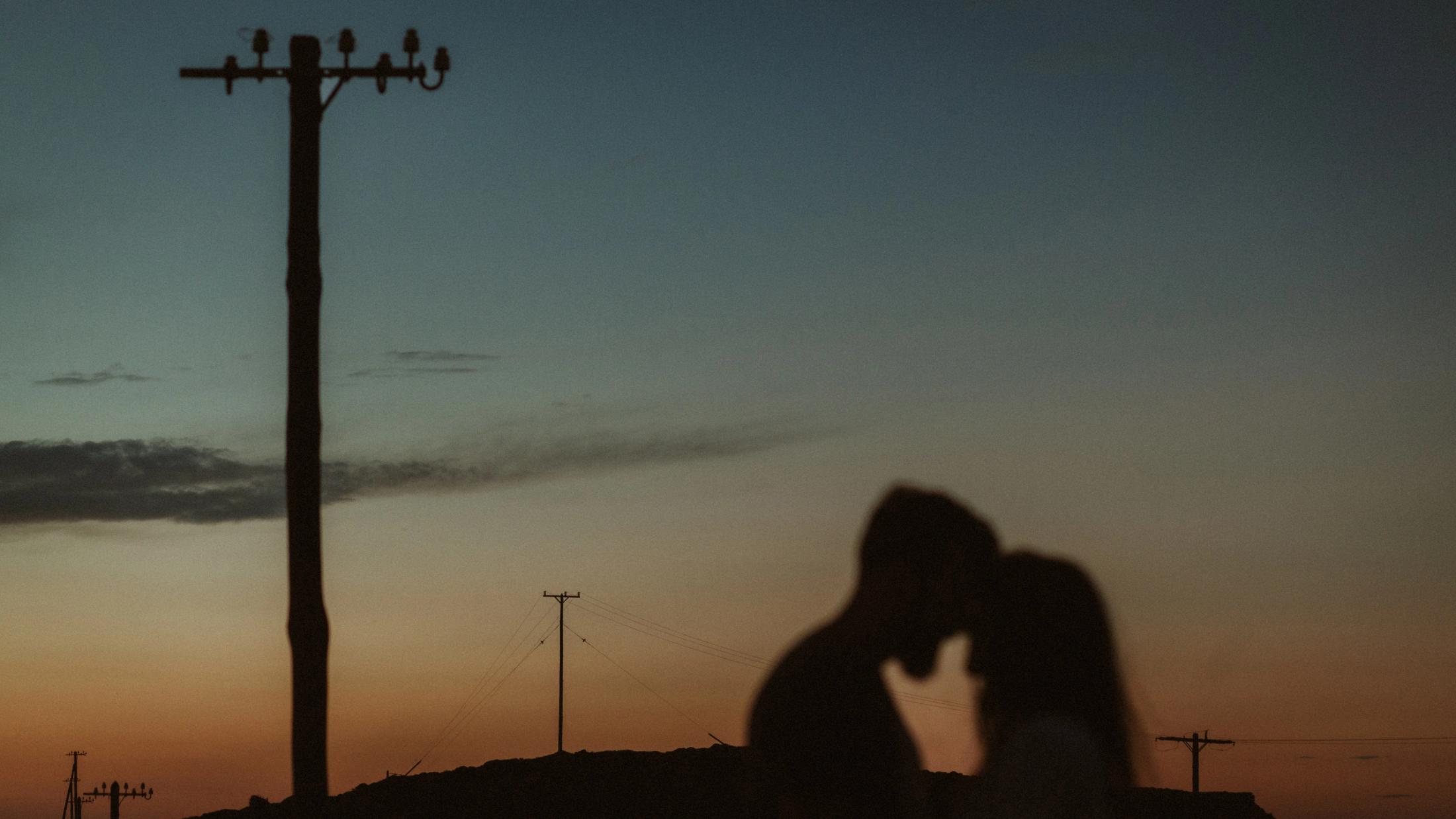 Eleni and George kiss as silhouettes beside a utility pole at sunset.