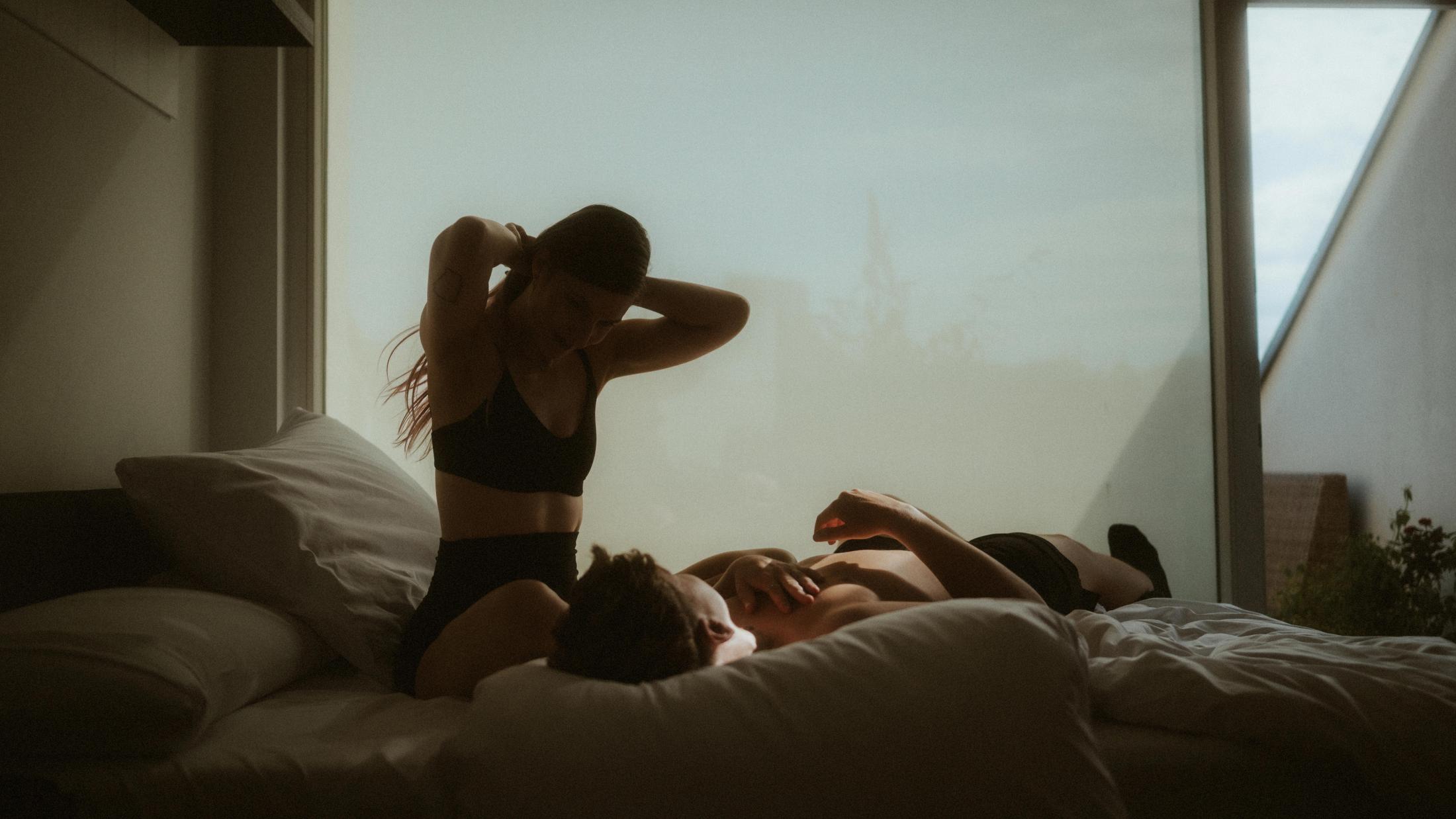 Wide view of Michèle sitting above Noah on a bed beside a large window.