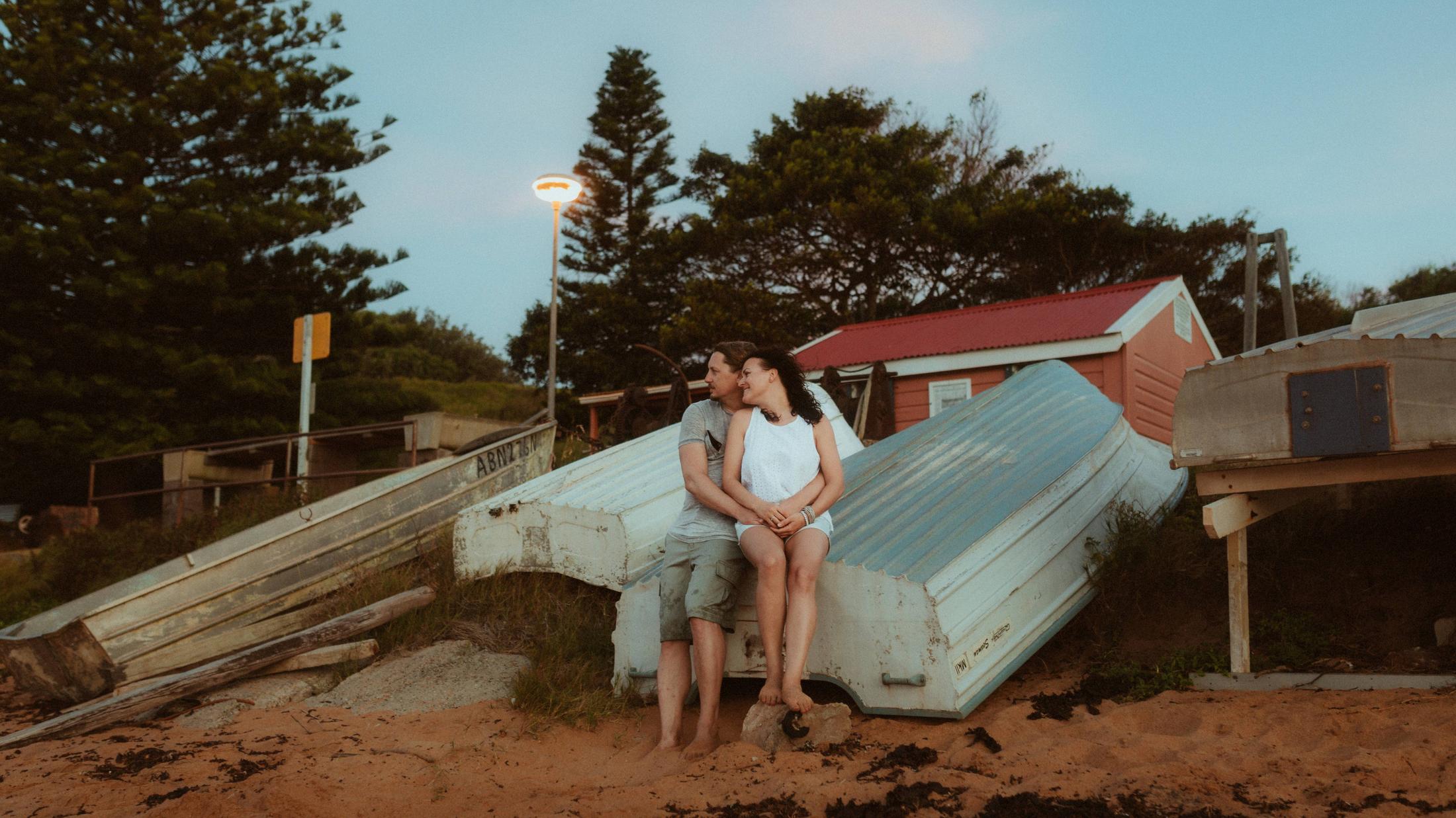Petra and Roman sitting beside overturned boats near a red boatshed