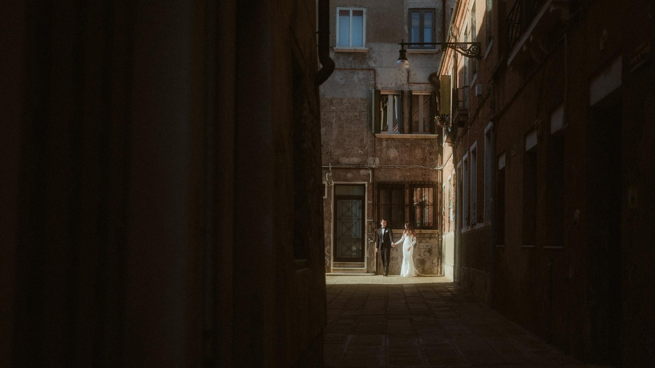 The couple appears as small figures at the bright end of a dark Venetian alley.
