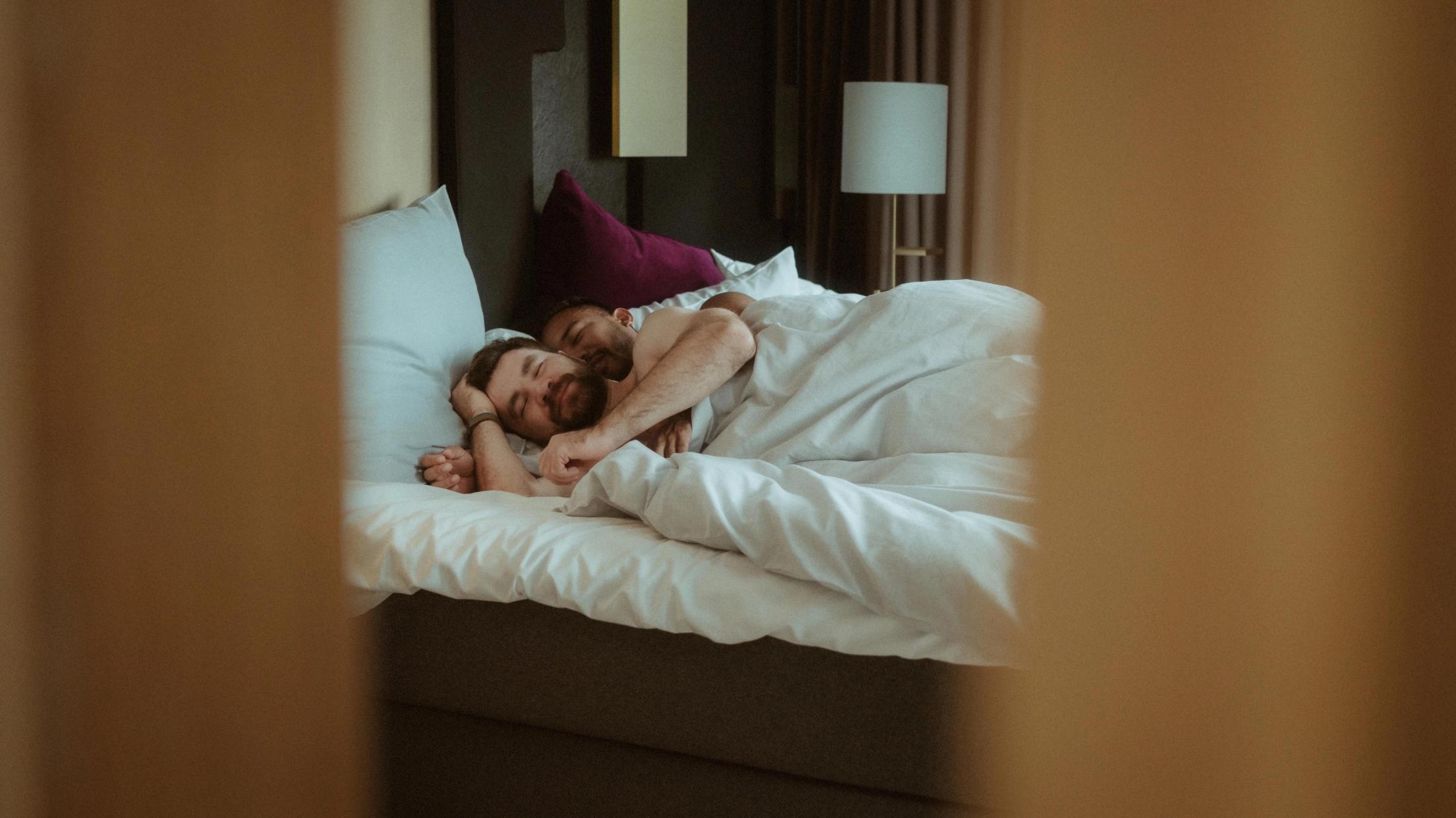 Wide view of Kris and Ilya resting together in bed inside their Copenhagen hotel room.
