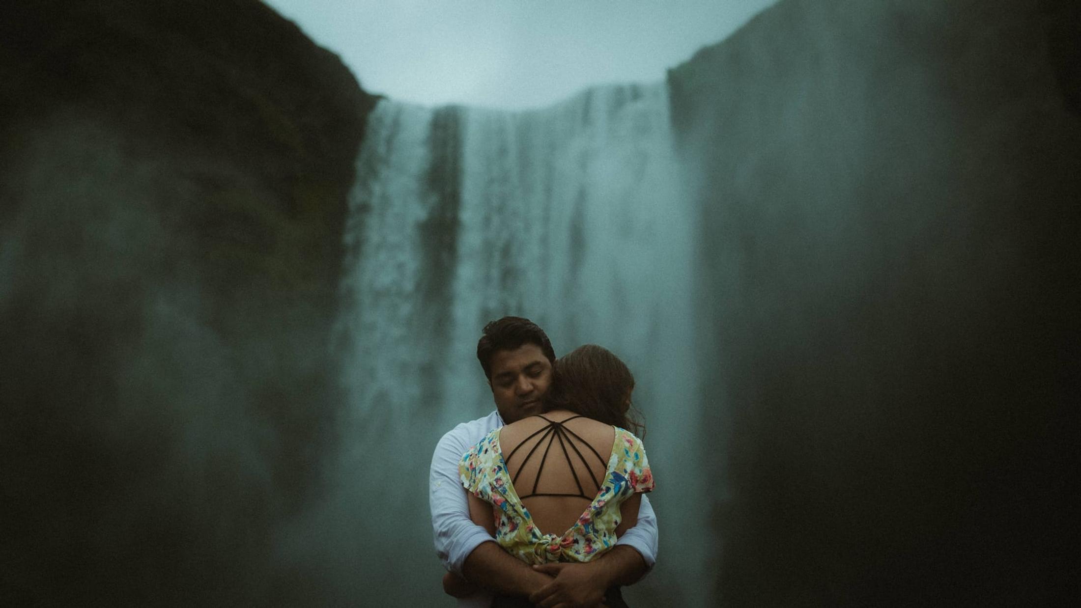Two people embracing with Skógafoss waterfall behind them.