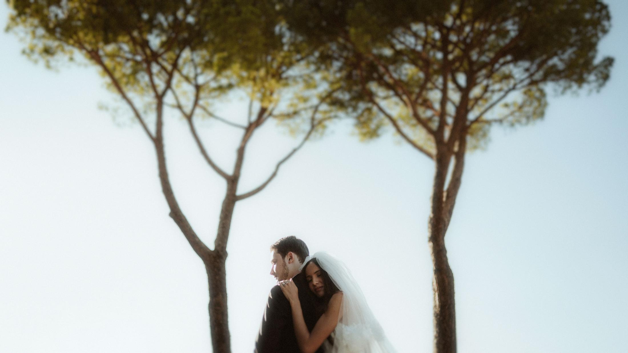 The couple embraces beneath a broad pine tree with a large pale sky above them.