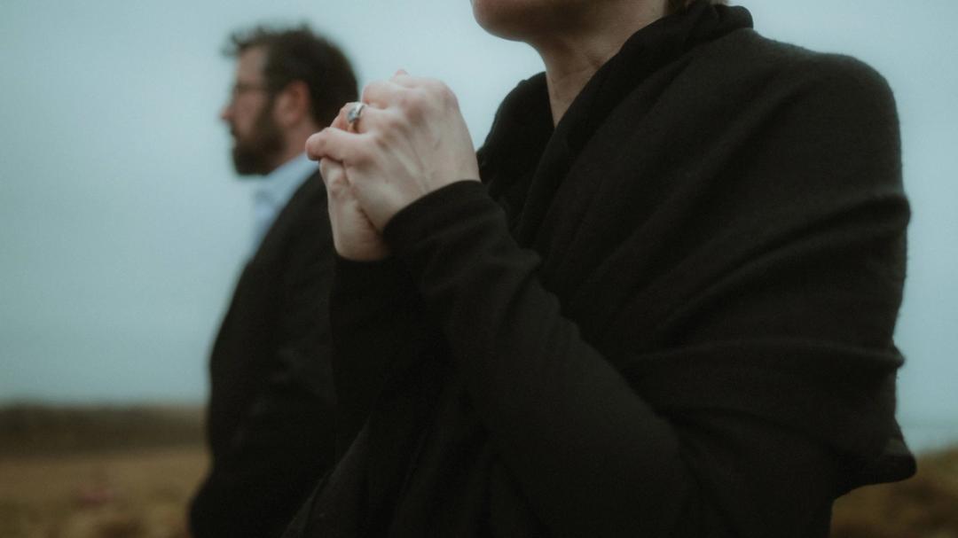 Gefaltete Hände liegen nahe einem vom Wind bewegten Gesicht, während eine zweite Person unscharf dahinter steht.