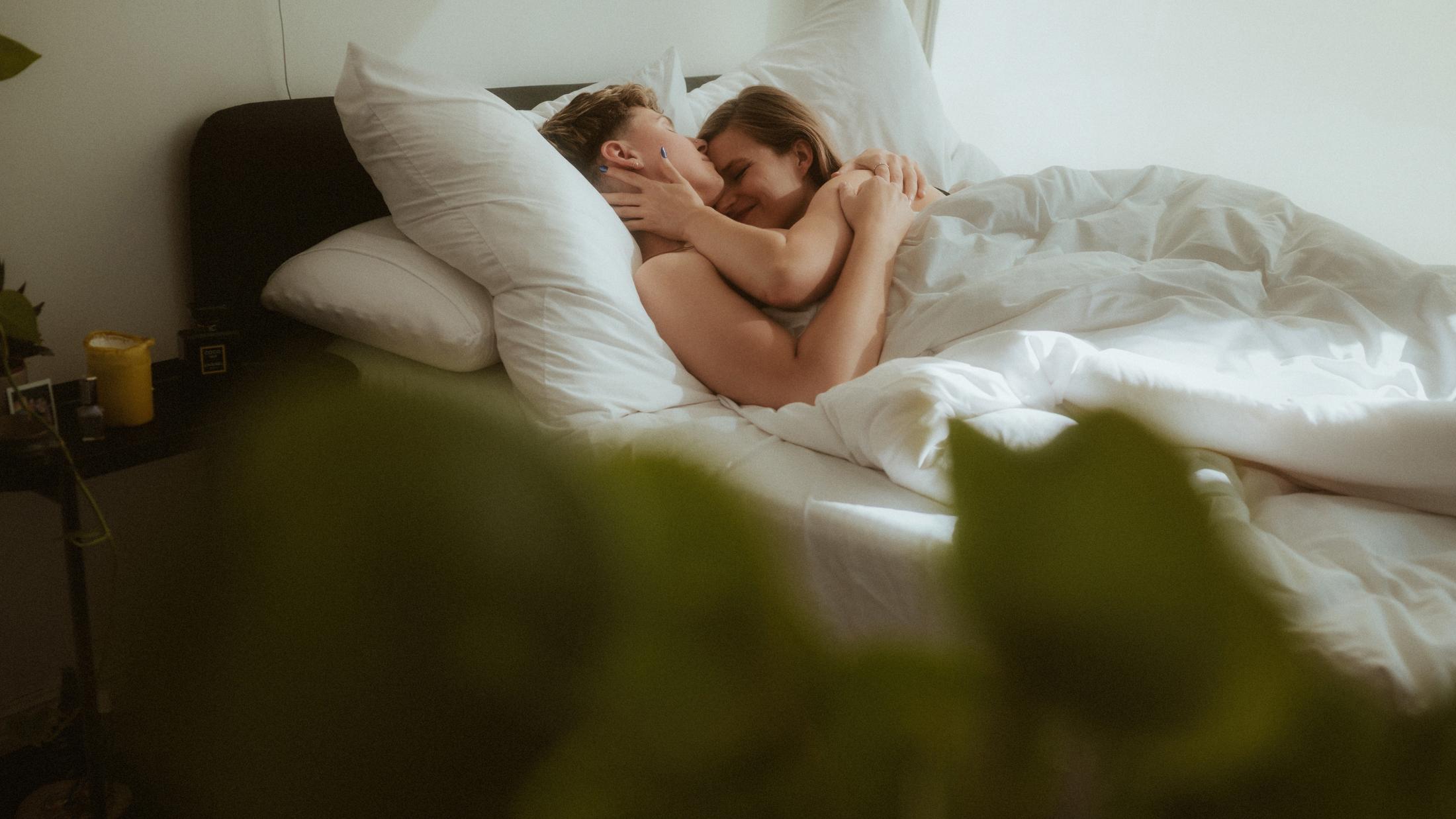 Noah and Michèle lying together beneath white sheets on a bright bed.