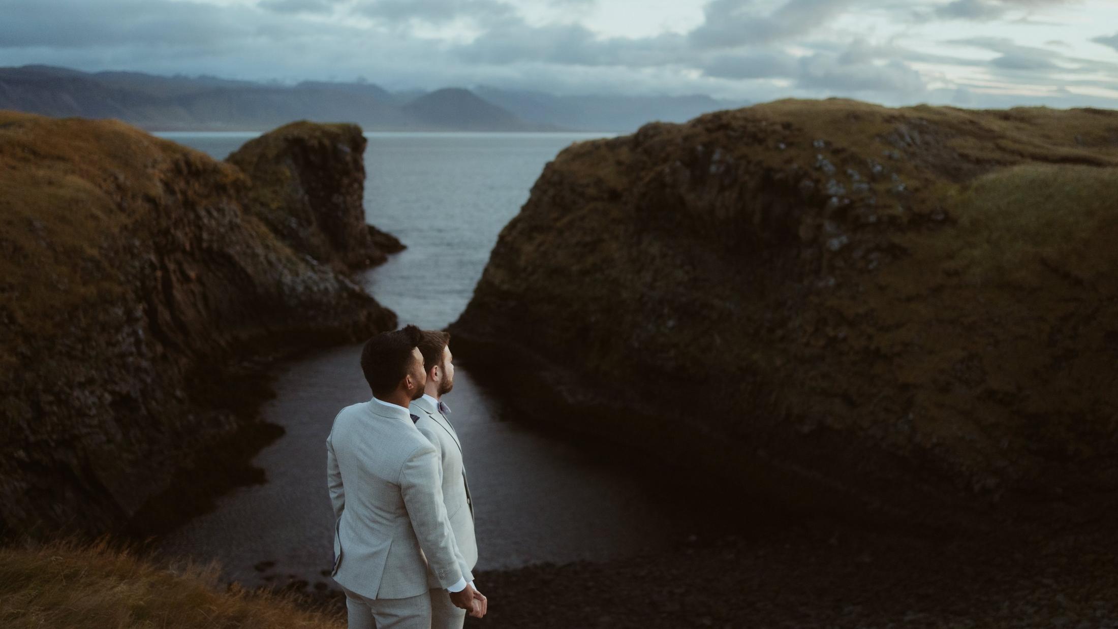 Two people close together above a rocky inlet and the sea.