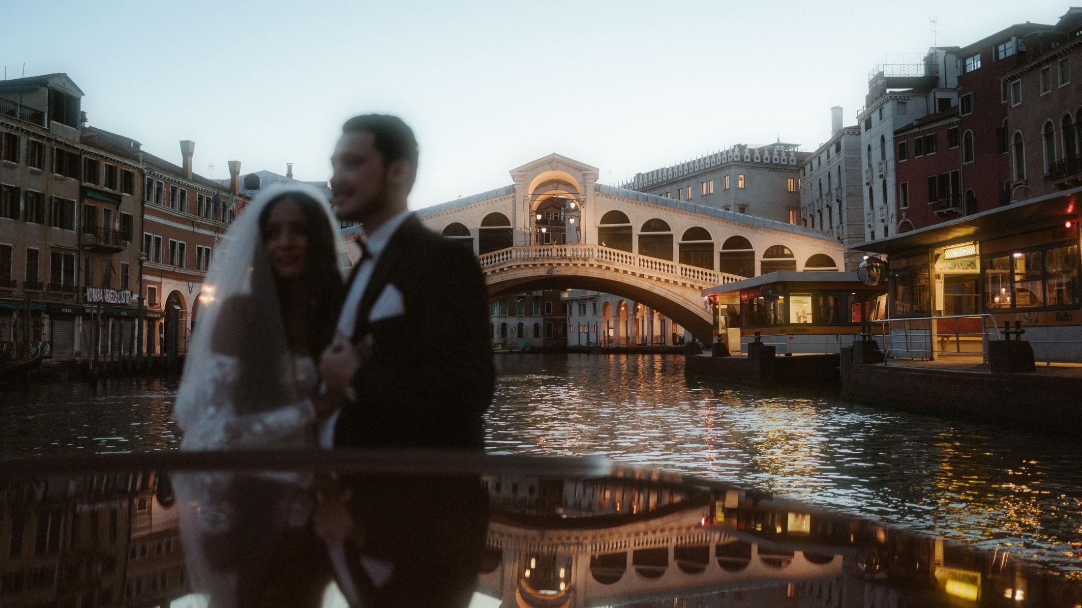 The couple appears softly blurred beside the Rialto Bridge and reflections on the water.