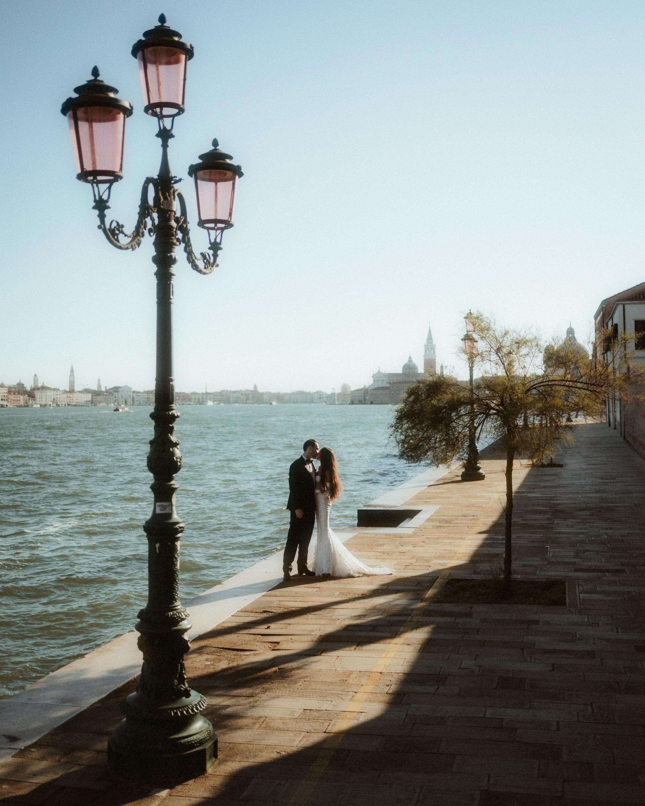 The couple embraces beneath a tall streetlamp beside the lagoon.